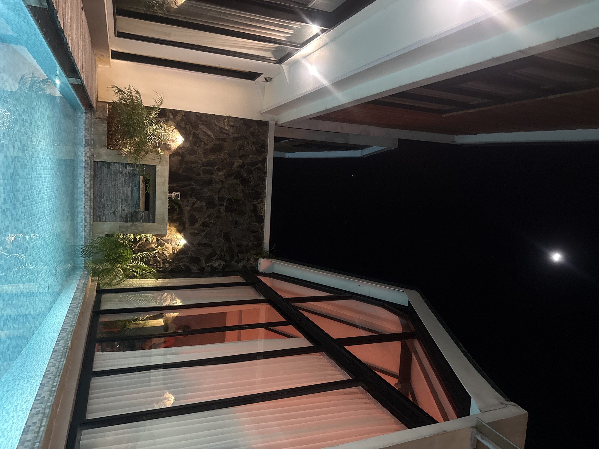 The image captures an architectural scene at night in Kediri, Indonesia. A swimming pool with light blue tiled walls occupies the left side of the frame. A wall of dark stone and vegetation borders the pool. Above is a partial view of a building structure with horizontal black and white lines. A second building overhangs the first and has visible wood paneling. The top right shows a dark night sky with a bright, singular light source resembling the moon. A triangular glass roof structure is visible at the lower center.