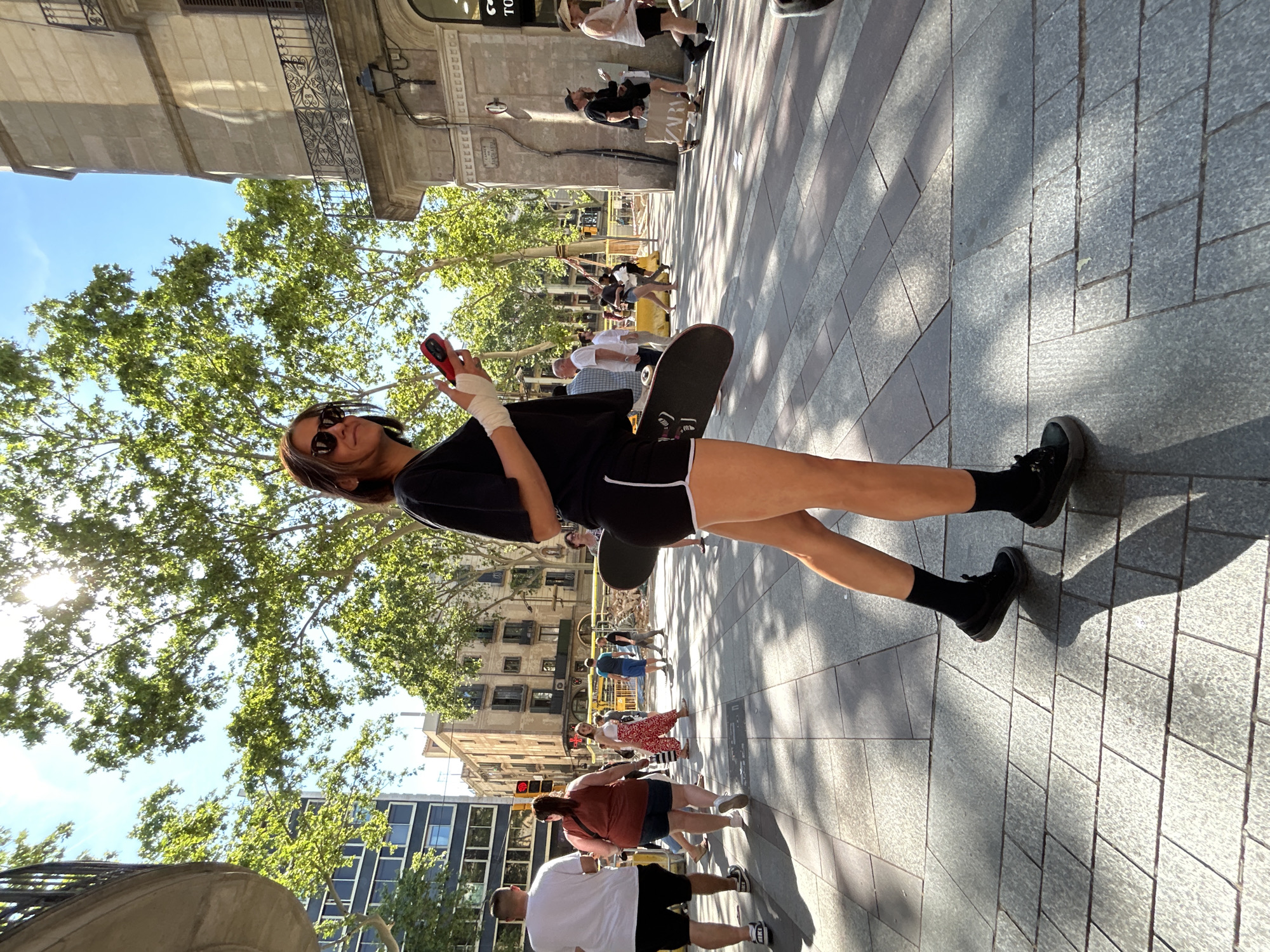 A young woman stands with a skateboard under her arm on a wide, pedestrianized street in Barcelona, Spain, holding a red cell phone in her right hand and looking towards the viewer with sunglasses on. She wears a black t-shirt, black shorts with white trim, black socks, and black shoes. Her right wrist is wrapped in a bandage. The street is paved with rectangular gray stones. Behind her are trees and buildings, including an ornate building with balconies. In the background, other pedestrians walk along the street.