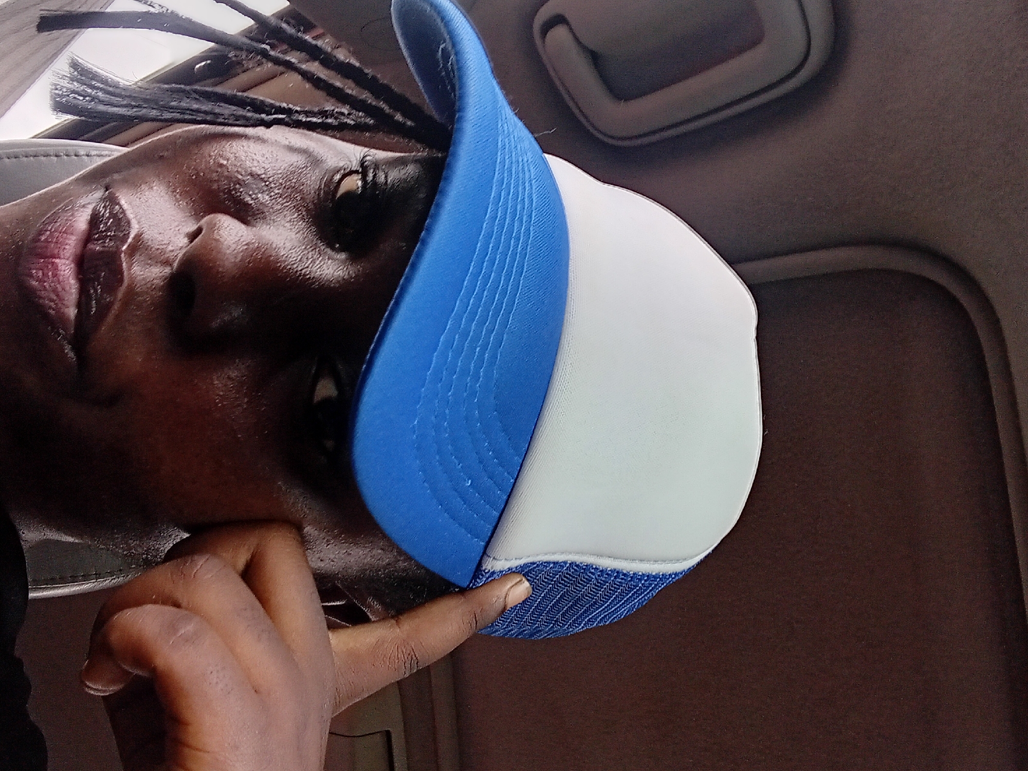 In Adoma, Nigeria, a close-up, low-angle selfie captures a person inside a vehicle. The subject is wearing a blue and white trucker hat and has braids. Their right index finger is touching the side of their face, near their temple, and their left eye is visible. The background consists of the vehicle's interior, including the headliner and a sun visor.