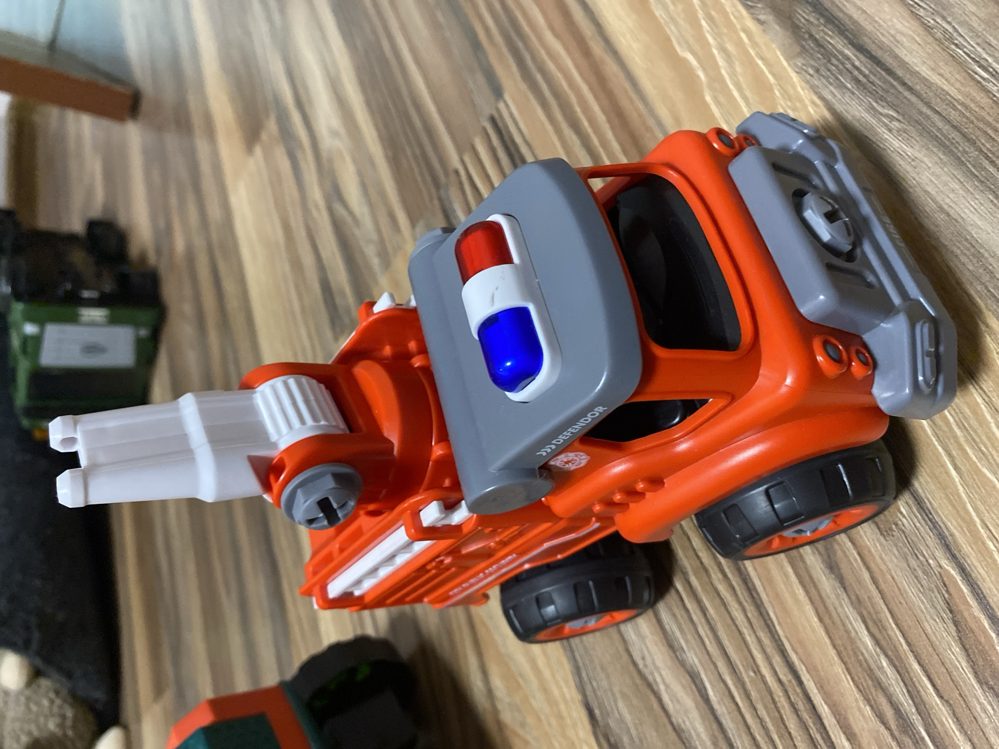 A close-up, high-angle shot shows a bright orange toy fire truck with grey accents, including a light bar with blue and red lights, parked on a wooden floor. The fire truck has large black tires with orange rims and a white ladder attachment. To the left, the green and orange front section of another toy truck is partially visible. In the background, a blurry dark object, possibly a stuffed animal, sits next to a green toy vehicle and a wooden table leg. The scene is indoors and does not depict any recognizable landmarks or details specific to Balashikha, Russia.