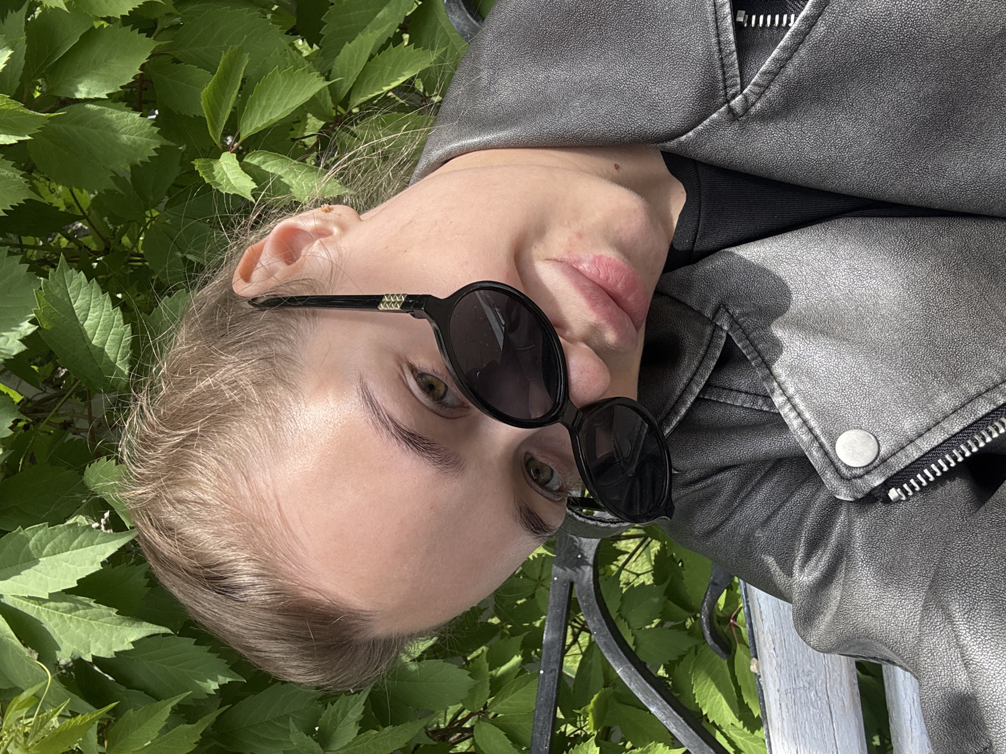 A woman is looking at the camera while sitting on a white wooden bench, surrounded by green foliage. She is wearing a black t-shirt under a silver leather jacket and dark sunglasses. The photograph appears to have been taken in an outdoor setting. No information is available from the image to indicate this location to be Chaikovsky, Russia.