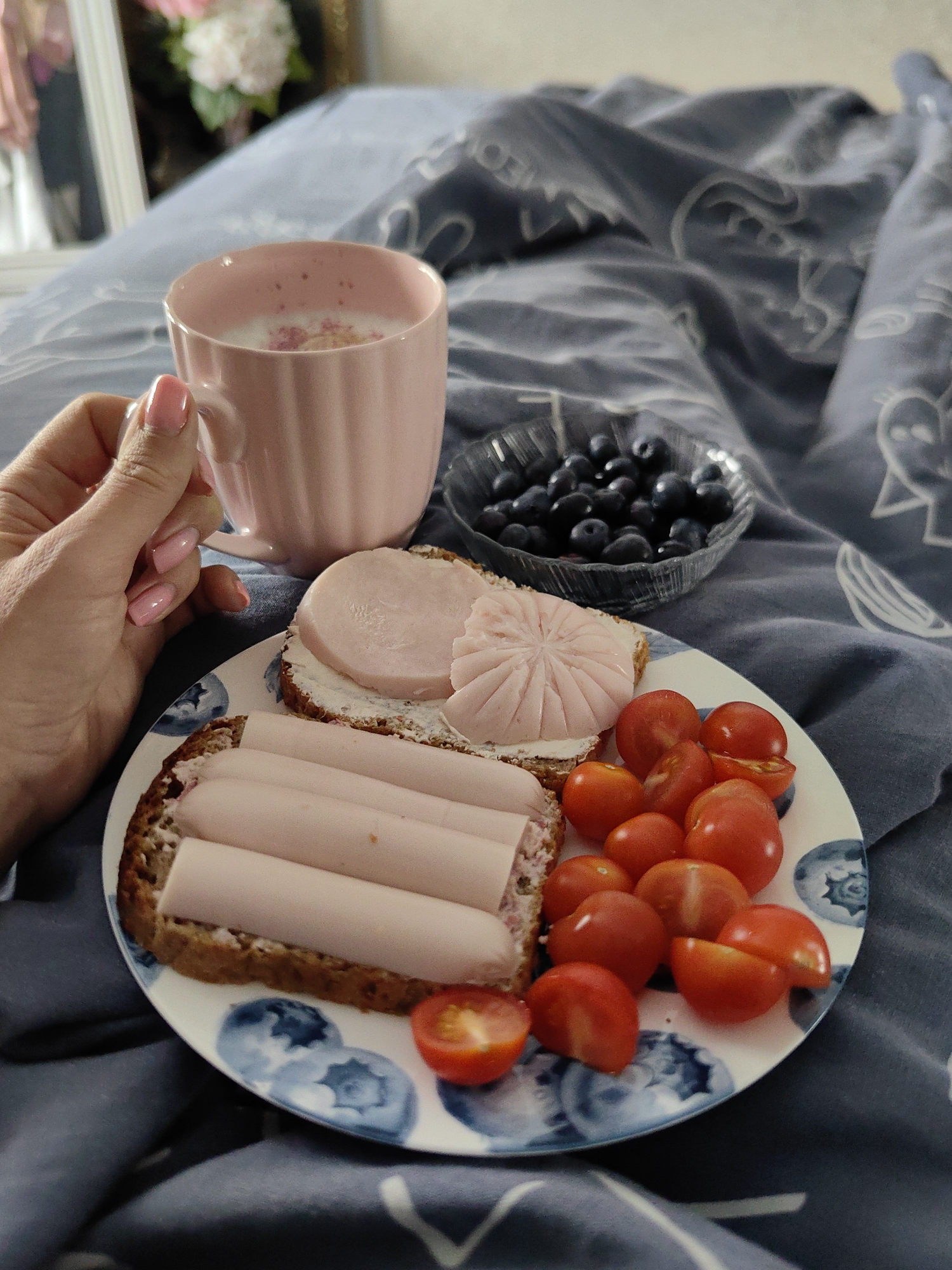 In Fanipal, Belarus, a hand with a pink manicure holds a pink mug filled with a pink beverage and flower petals. Beside it, a small glass bowl overflows with blueberries. A plate displays two open-faced sandwiches topped with cheese and sliced ham, accompanied by a cluster of halved cherry tomatoes. The scene is set against a backdrop of blue bedding with a pattern of white figures.