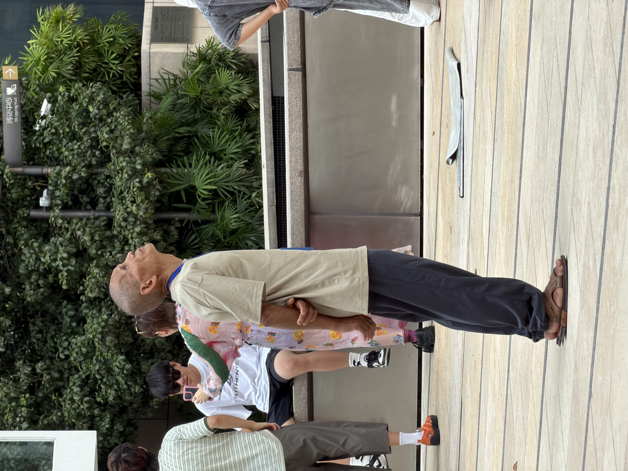 In Yuzhong District, China, an elderly man with a bald head stands facing right, looking upwards. He wears a light beige t-shirt and dark grey pants, with brown sandals on his feet. His hands are clasped behind his back. Behind him, a person wearing a pink floral dress stands and another person in a white t-shirt with black shorts and sunglasses holds up a pink mobile phone. To the far left, a person in a green and white striped t-shirt and grey pants stands. The background consists of lush green foliage and a grey concrete structure. A sign pointing right indicates "Shopping Mall". The ground is paved with light-colored tiles.