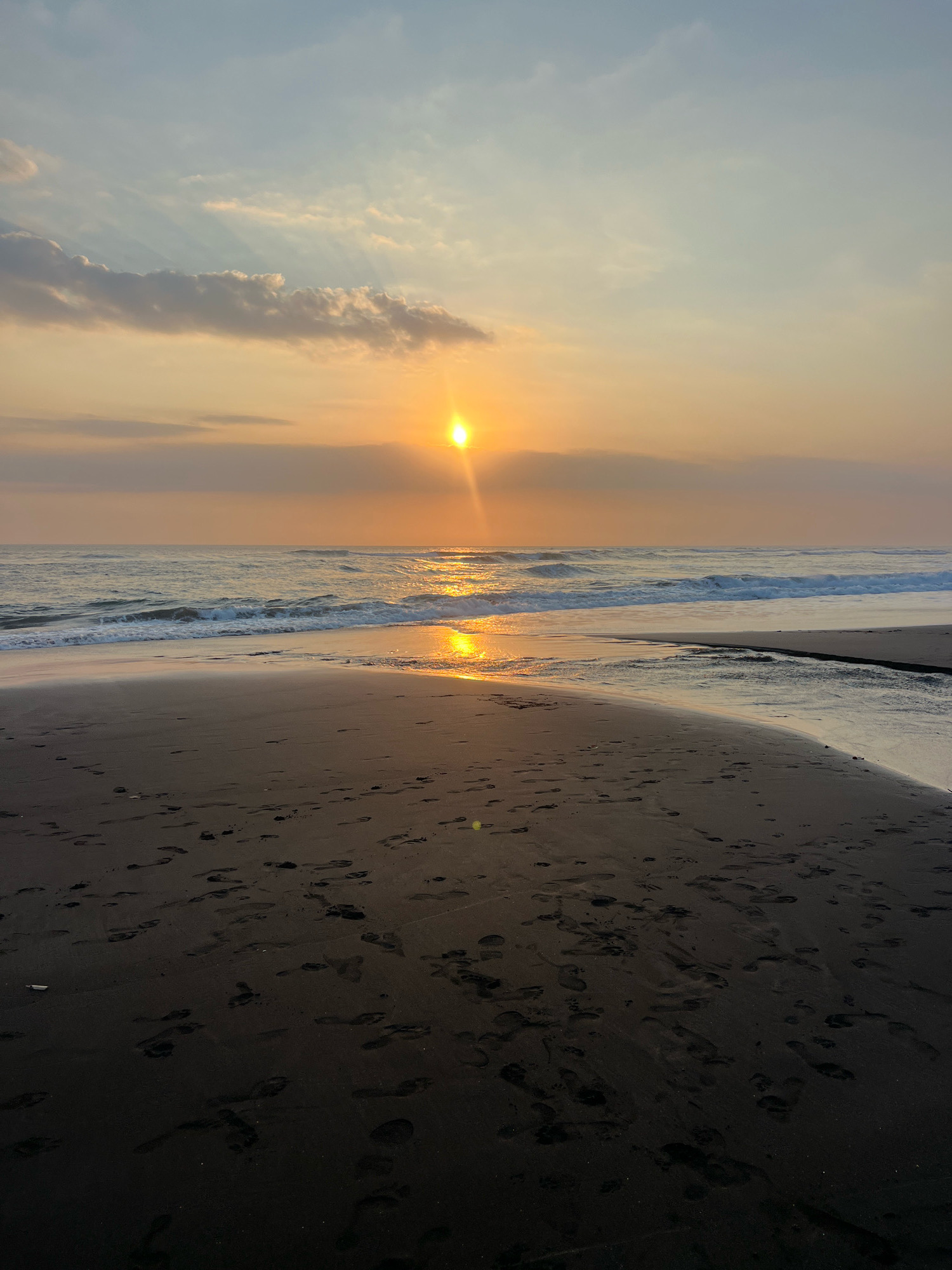 This is a photograph of a beach at sunset. The sun is low on the horizon, casting a warm, golden glow over the scene. The sky is a gradient of pale blue and orange, with a few wispy clouds. Gentle waves are rolling in from the ocean, creating a foamy white surf. The sand is dark and wet near the water's edge, reflecting the sunlight. Footprints are visible in the sand, indicating recent human activity. The overall atmosphere is serene and peaceful. There is no identifiable information to suggest this location is in Kediri, Indonesia.