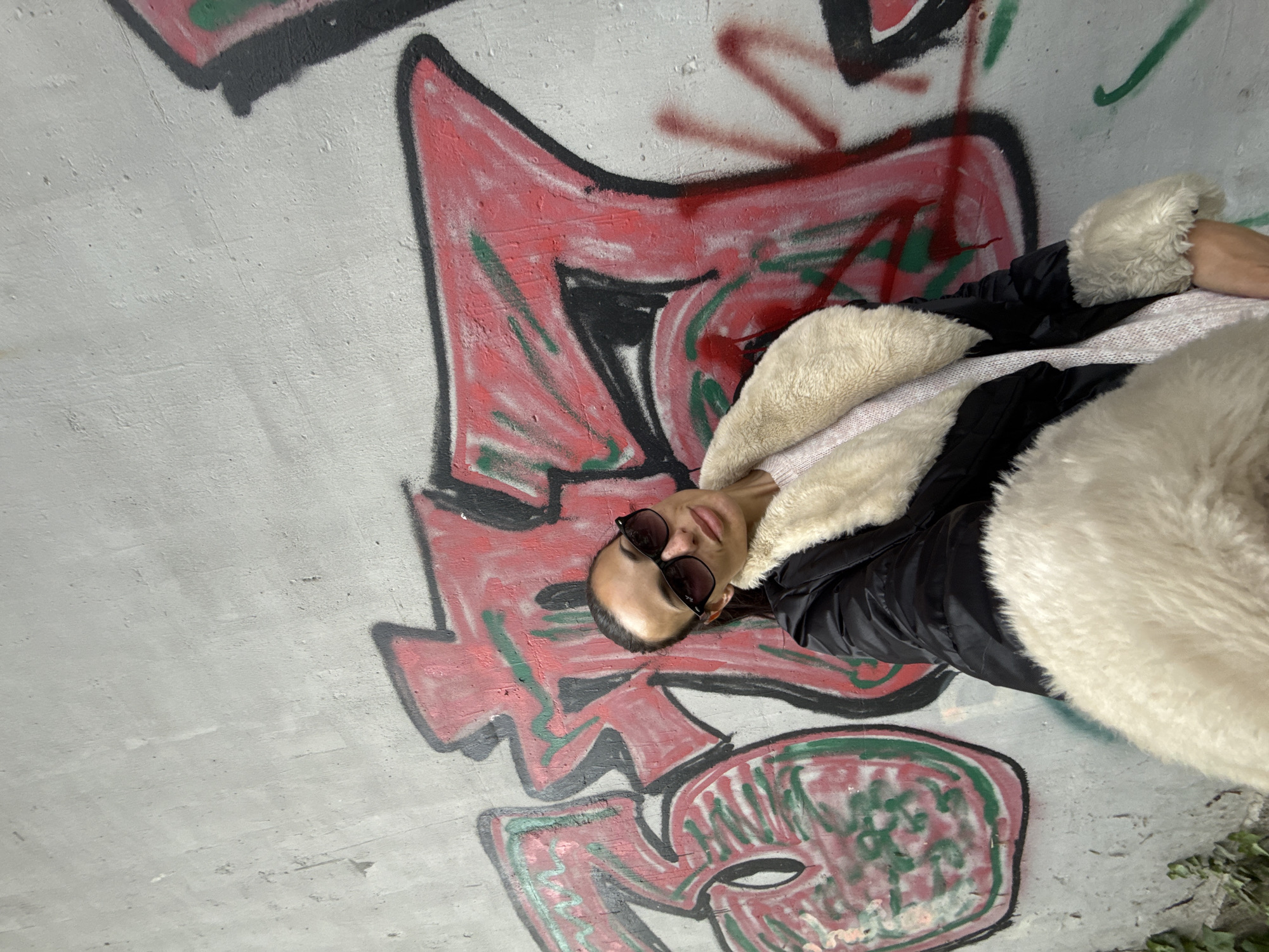 An individual, likely female, is depicted lying or leaning horizontally against a graffiti-covered concrete wall. She has dark hair pulled back and wears dark sunglasses pushed up onto her head. She is dressed in a black puffer jacket that features a prominent, plush cream or off-white faux fur lining or trim, possibly indicating a full faux fur coat. A light pink or cream-colored knitted garment is visible underneath.
The background is a textured, light grey concrete wall heavily adorned with vibrant graffiti. The graffiti primarily consists of large, stylized shapes and letters outlined in black, filled predominantly with red, and accented with patches of green. While the designs suggest urban tagging or street art, no specific words are clearly legible from the visible sections. The wall's surface shows signs of age and wear, consistent with an outdoor urban setting like an underpass or a neglected building exterior in Pleven, Bulgaria.
The activity appears to be a posed photograph, with the subject looking upwards towards the camera with a relaxed expression. The lighting suggests it is daytime, possibly overcast or within a shaded area, as there are no strong shadows. The attire implies cooler weather. No explicit text is discernible within the graffiti.