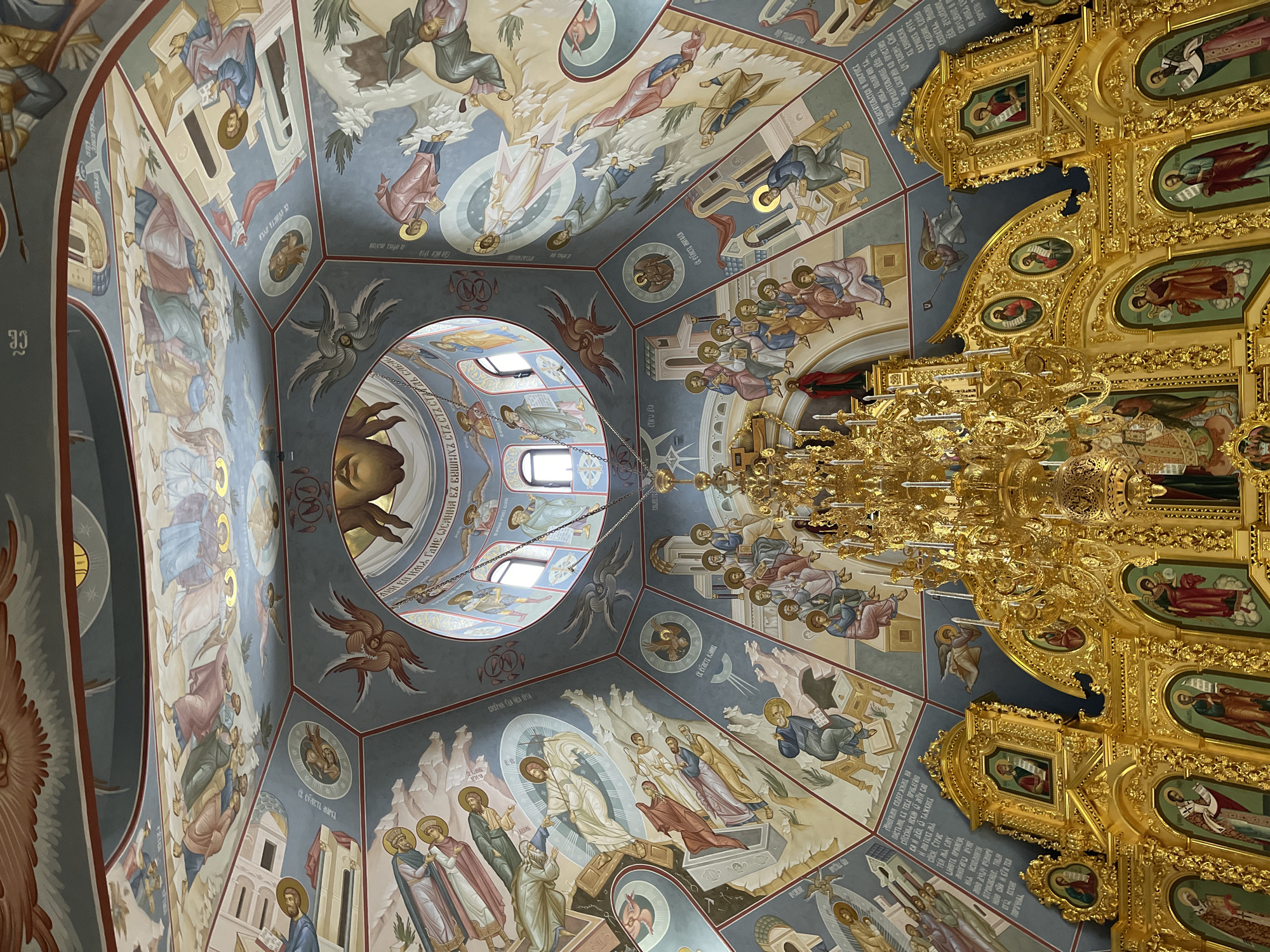 The image captures an interior view of a church or religious building, likely in Khimki, Russia, as indicated by the presence of Cyrillic text. The primary focus is on the ceiling, which is elaborately decorated with religious frescos, painted in an Orthodox Christian style. These frescos depict various religious figures, scenes, and symbols.
A large, ornate, golden chandelier hangs from the ceiling, partially obscuring some of the frescos. A circular opening in the ceiling allows natural light to enter, illuminating the interior.
Part of a golden iconostasis, a wall of icons and religious paintings, is visible on the right side of the image. The overall environment suggests a place of worship, characterized by intricate religious art and architectural details.