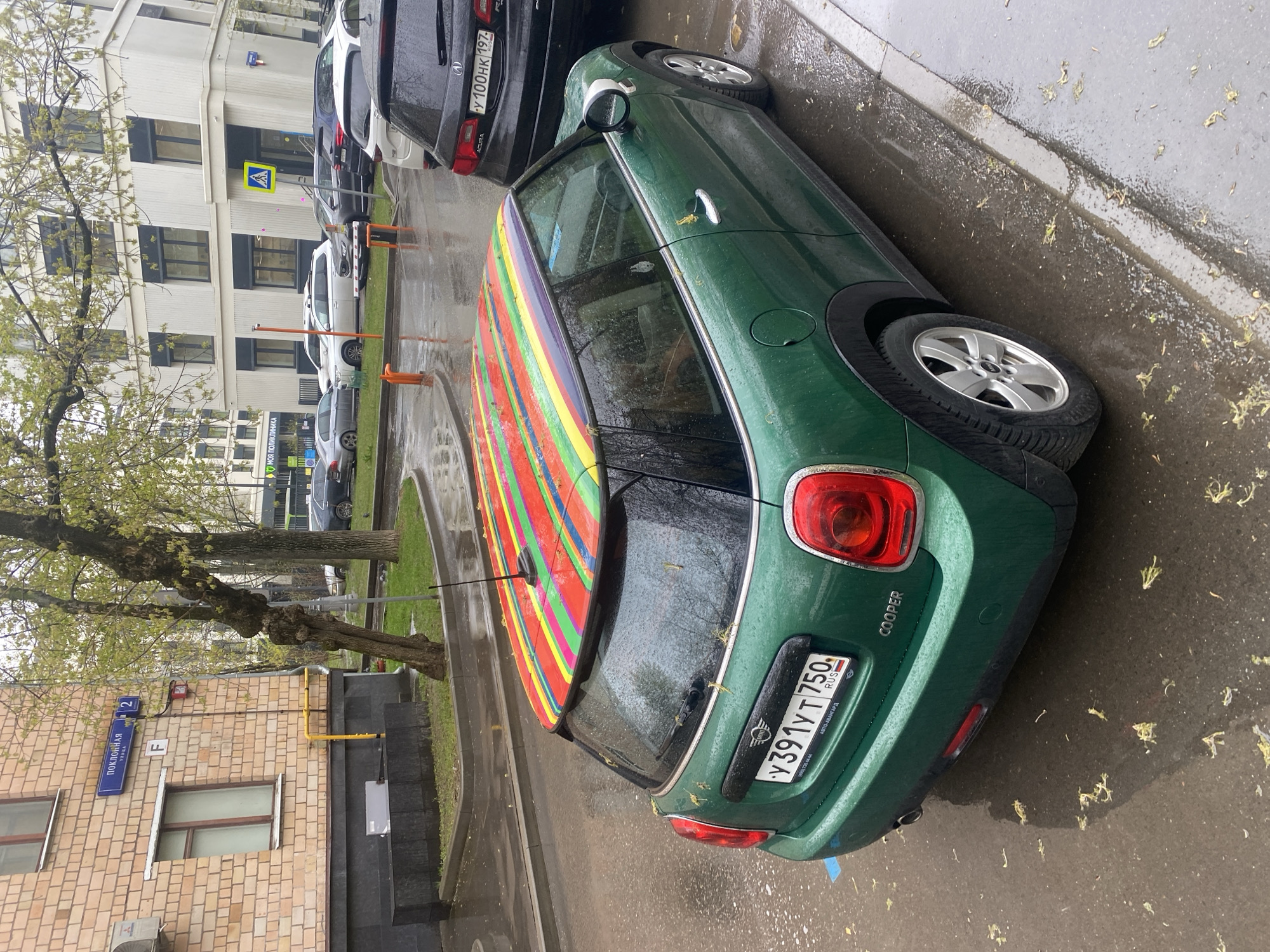 The image shows a green Mini Cooper with a multicolored striped roof parked on a wet street in Moscow, Russia. The car's license plate reads "Y391YT750 RUS." A street sign on a nearby building indicates the location is "Поклонная улица 2." Other parked vehicles are visible in the background, along with a multi-story building and a tree. The weather appears to be rainy or recently wet.