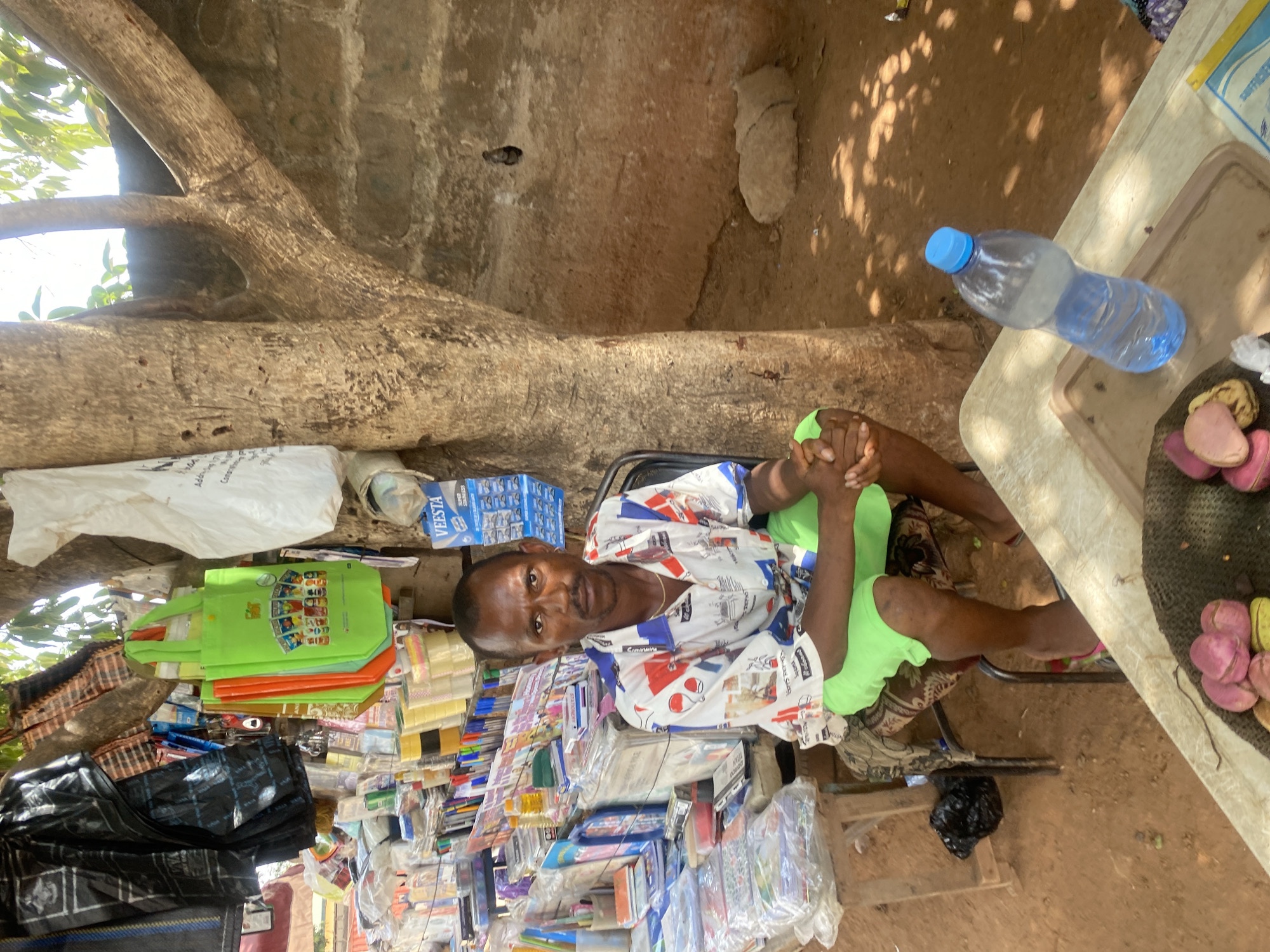 This image depicts an outdoor vendor stall in Sabon Tasha, Nigeria. One adult male is seated, facing directly forward, positioned left of center. He wears a short-sleeved white collared shirt with red, blue, and black geometric and abstract patterns, and bright green shorts. His hands are clasped in front of him. He has short dark hair, a light mustache, a short beard, and small hoop earrings in both ears. A gold-colored chain is visible around his neck. He sits on a dark-framed chair with a patterned fabric seat.
To the left of the seated man, a display of assorted merchandise is arranged on a wooden table and shelving units. This includes numerous packaged notebooks, pens, markers, clear and yellow adhesive tapes, and various small accessories. Overhead, black and white plastic bags hang from a tree branch or structure, alongside several bright green reusable shopping bags depicting cartoon characters. A blue display card for "VEESTA SUPER STAINLESS" razor blades is affixed to a tree.
In the lower right foreground, a light-colored rectangular table holds a brown plastic tray. On the tray, a clear plastic bottle with a blue cap is approximately half-full of water. Adjacent to the bottle, a dark woven basket or mat contains several kola nuts; some are peeled, displaying a reddish-pink color, while others retain their greenish-yellow husks.
The ground is composed of reddish-brown earth, showing patterns of dappled sunlight. A large, light-brown tree trunk rises to the right of the seated man, with branches extending upwards and right. A light-colored, rough-textured wall, constructed of concrete or mud bricks, forms the background on the right side of the scene. The upper portion of the image reveals bright daylight and some green foliage.