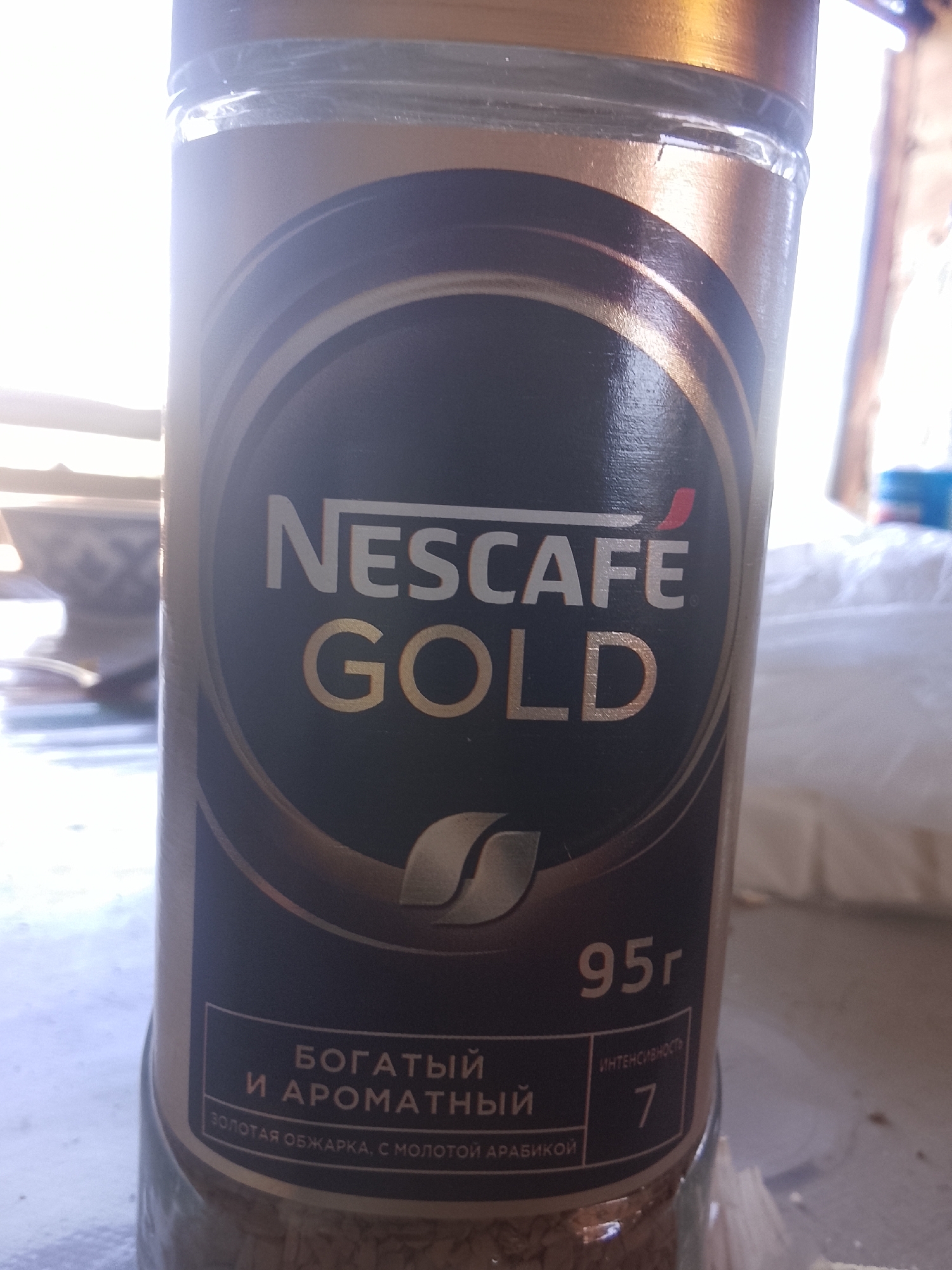 The image shows a close-up of a glass jar of Nescafé Gold coffee. The jar is predominantly dark brown with gold accents. The Nescafé Gold logo is prominently displayed in white and gold lettering. Below the logo, in Russian, it reads "БОГАТЫЙ И АРОМАТНЫЙ" (Rich and Aromatic) and "ЗОЛОТАЯ ОБЖАРКА, С МОЛОТОЙ АРАБИКОЙ" (Golden Roast, with Ground Arabica). To the right, it indicates "95г" (95g) and an intensity rating of "7" labeled "ИНТЕНСИВНОСТЬ" (Intensity). The background is blurred, but suggests an indoor setting with natural light coming from the right, casting a bright glow. There is no discernible indication of the city of Sabir Rakhimov, Uzbekistan, in the image.