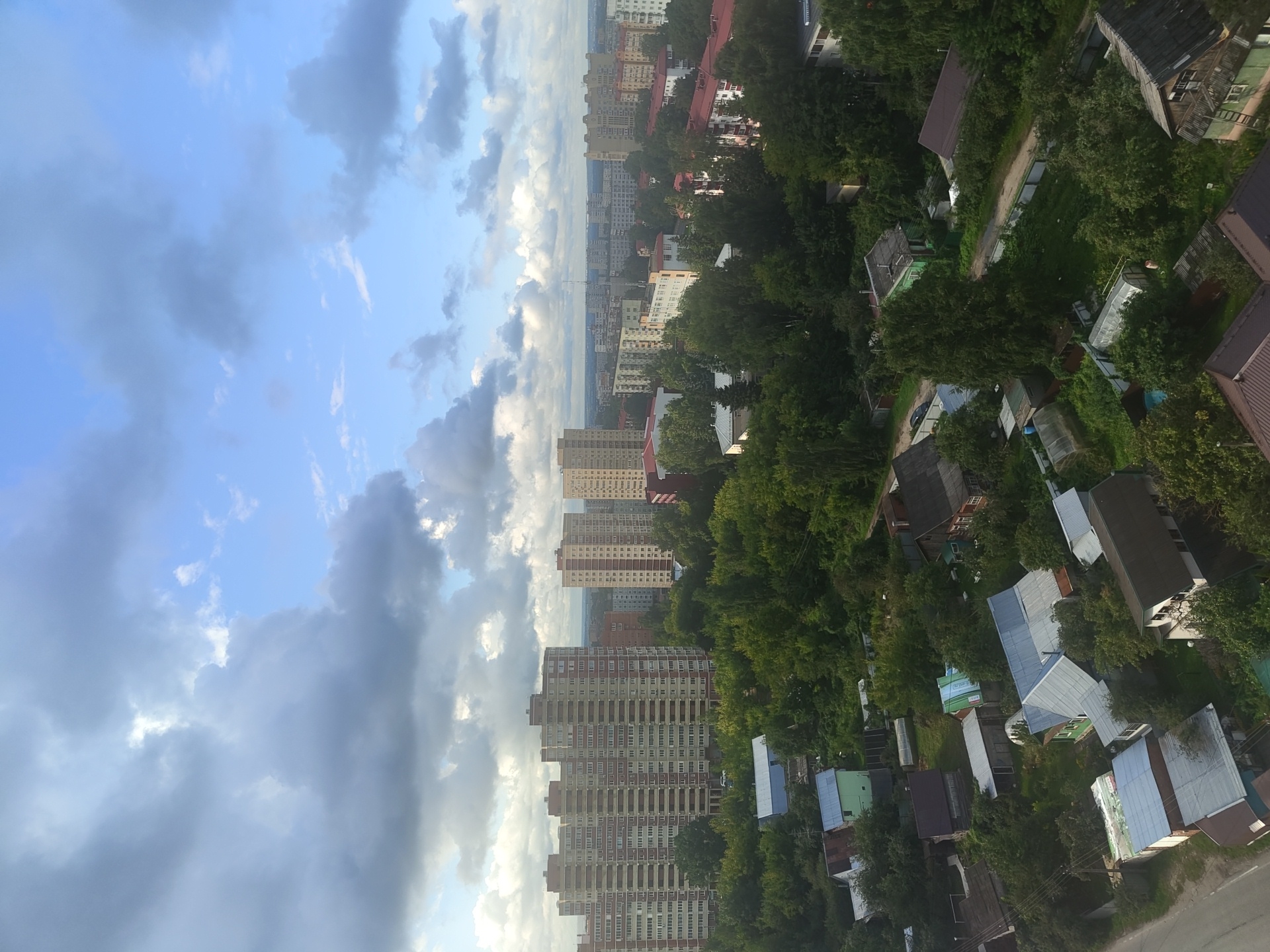 This is an aerial, high-angle, daylight shot of the city of Ramenskoye, Russia. The sky is partly cloudy with dramatic grey clouds mixed with patches of blue sky. In the foreground, a dense canopy of green trees dominates the lower half of the image, interspersed with various residential structures. These include numerous low-rise houses with pitched roofs, some appearing to be older wooden structures with some with metal roofing, and several greenhouses. A road is visible on the left side of the frame. Further back, the landscape transitions to taller, multi-story apartment buildings, characteristic of Soviet-era urban development. These apartment blocks are a mix of beige, red, and white exteriors with numerous windows. Some of these buildings are quite large, stretching horizontally across the frame. In the distance, the cityscape continues with more buildings and a hazy horizon line, suggesting a moderately sized urban area. No people or vehicles are clearly discernible in the scene. The overall impression is of a residential area with a mix of older, detached housing and more modern, high-rise apartment complexes surrounded by lush greenery.