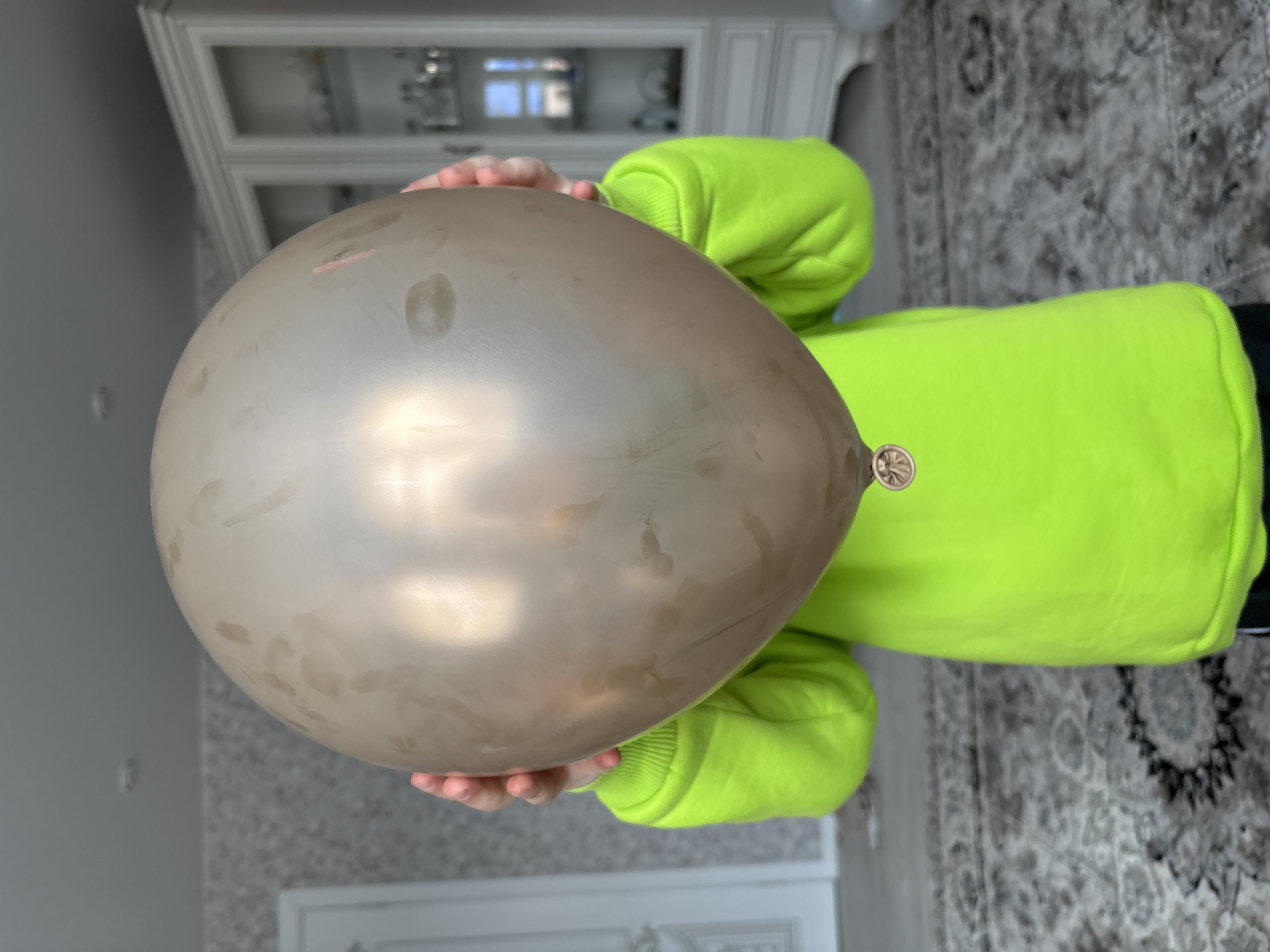 A child wearing a neon green sweatshirt is holding a dirty brown balloon in front of their face. The background is a room with patterned wallpaper, a white cabinet, and a patterned rug. There is a door in the lower background. The photo was taken indoors. The location is not discernible from the provided image.