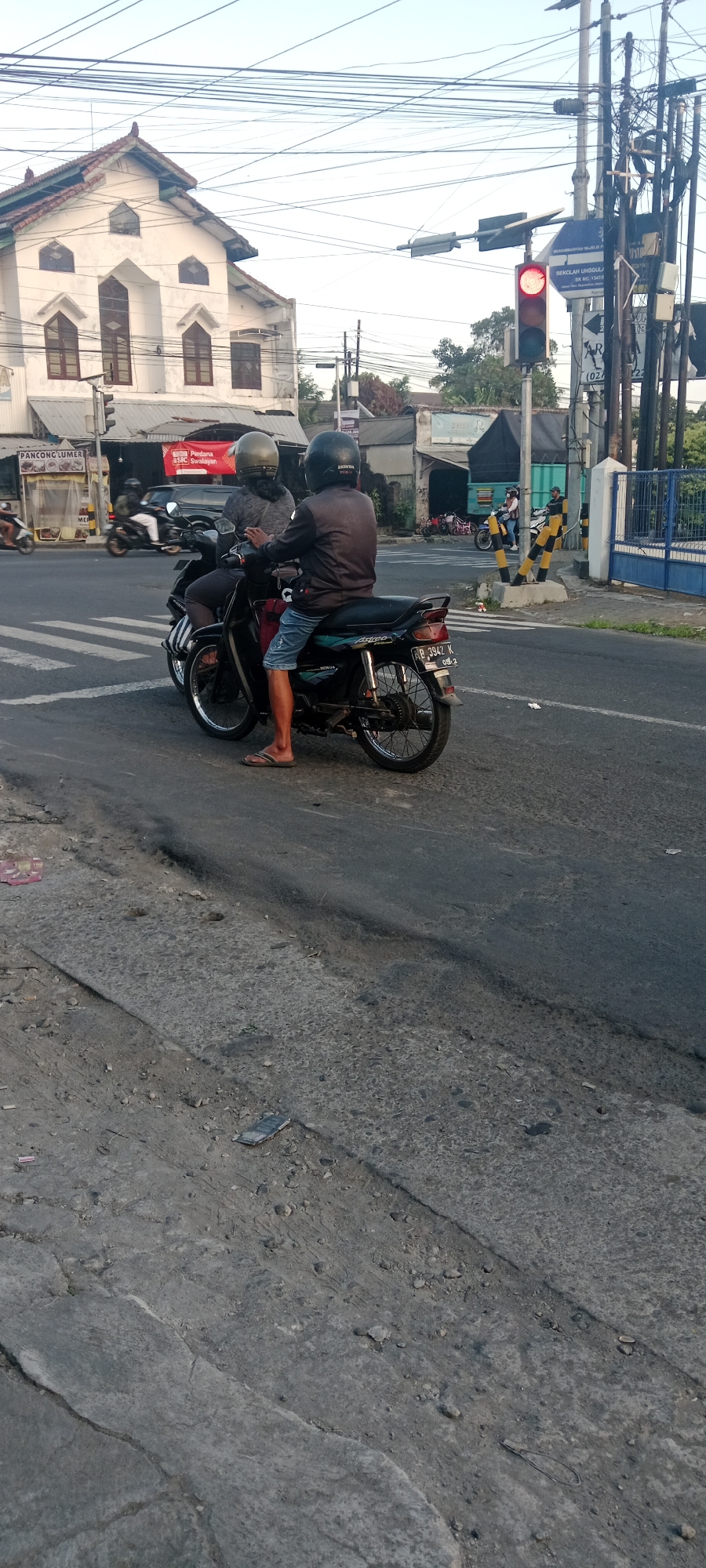 The image captures a street scene in Maguwoharjo, Indonesia, with a focus on two individuals on a motorcycle stopped at an intersection with a red traffic light. The motorcycle, a black Honda Astrea Grand with license plate B 3942 N, is positioned on a pedestrian crossing. The rider in the front wears a dark jacket and blue shorts, while the passenger behind wears a grey top. Both are wearing helmets. In the background, a white building with a distinctive arched facade is visible, possibly a place of worship or a school, with signage for "Pondok Pesantren Perdana" and "Sekolah Unggulan". The street is paved with asphalt and shows signs of wear and tear, with some debris on the ground. Overhead power lines crisscross the sky. The traffic light is red, indicating a stop. To the right, a blue and white gate and more electrical poles are present. The overall lighting suggests it is late afternoon or early morning.
