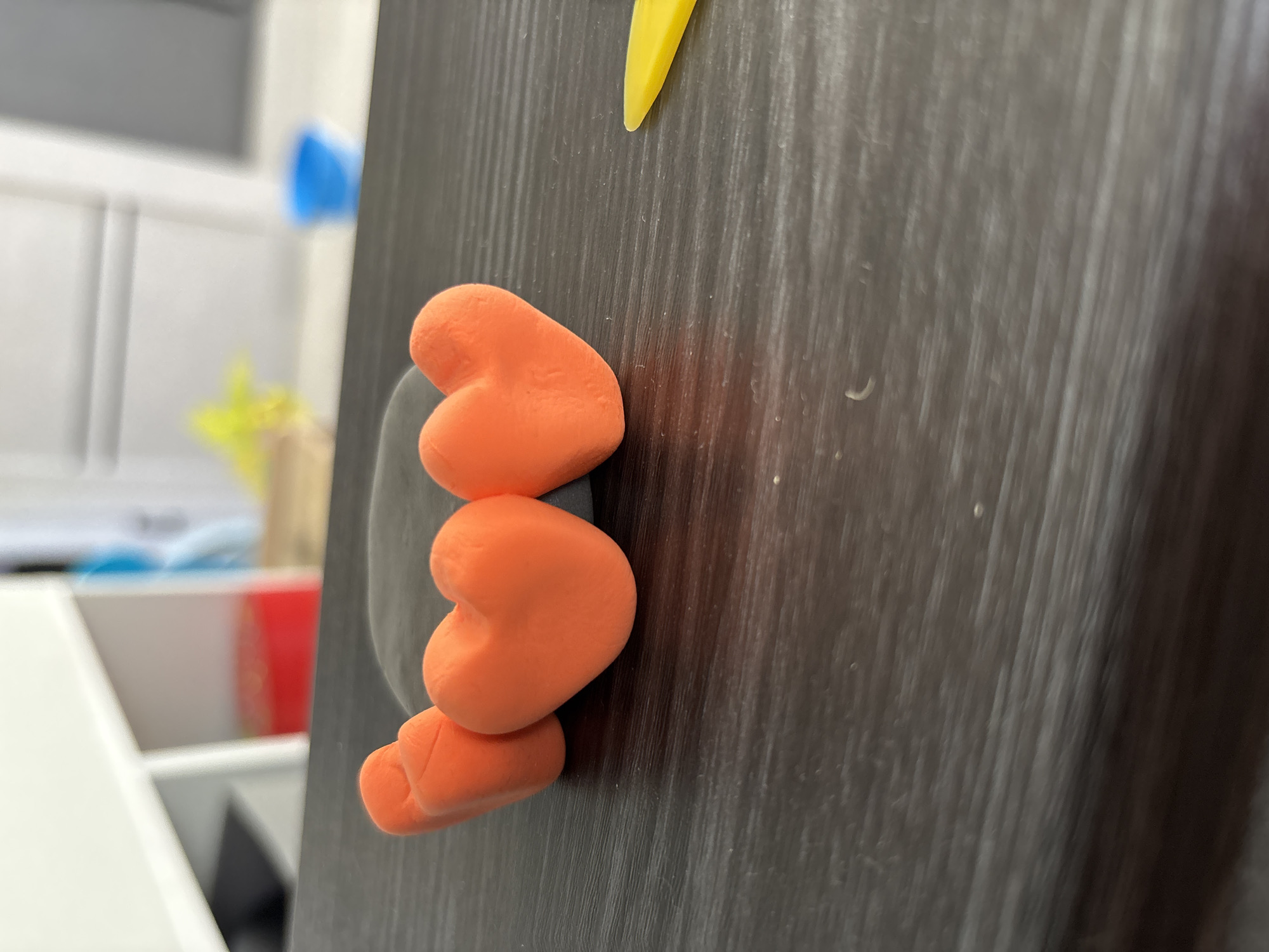 The image shows a dark, vertically striped wooden surface with three orange, heart-shaped clay figures stacked on top of each other. A small piece of yellow clay is visible at the top. The background shows a blurred domestic scene, including a white cabinet, a blue object, and a yellow object. The location cannot be determined from the image, so the request for "location in the city of Chaikovsky, Russia" is unfulfilled.