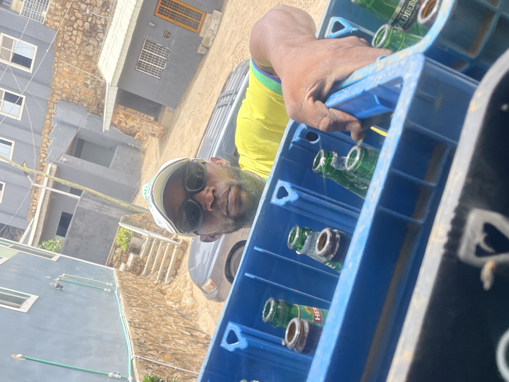 The image shows a man carrying a blue crate of beer bottles, likely in Gura Top, Nigeria. The man wears a yellow shirt, sunglasses, and a white cap. The beer bottles are green and appear to be "Goldberg Lager Beer". In the background, there are buildings with gray walls and a stone base, suggesting a residential or commercial area. A silver vehicle is also visible.
