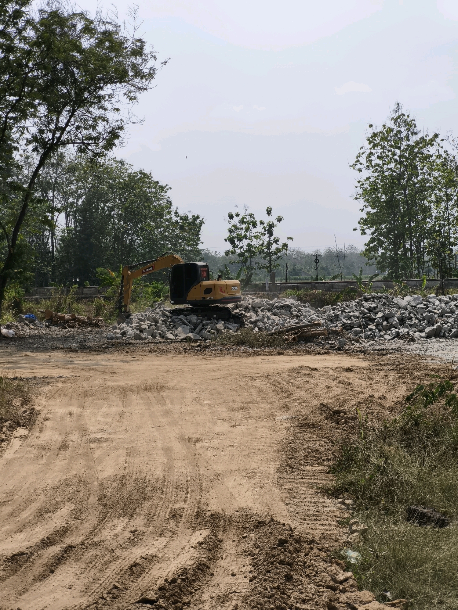 In Nambangan, Indonesia, an excavator is actively engaged in construction work. The yellow and black heavy machinery is situated amidst a large pile of gray rocks and debris, with its arm extended and bucket positioned over the stones. The surrounding environment is a construction site featuring a dirt road with tire tracks, vegetation, trees, and a hazy sky.