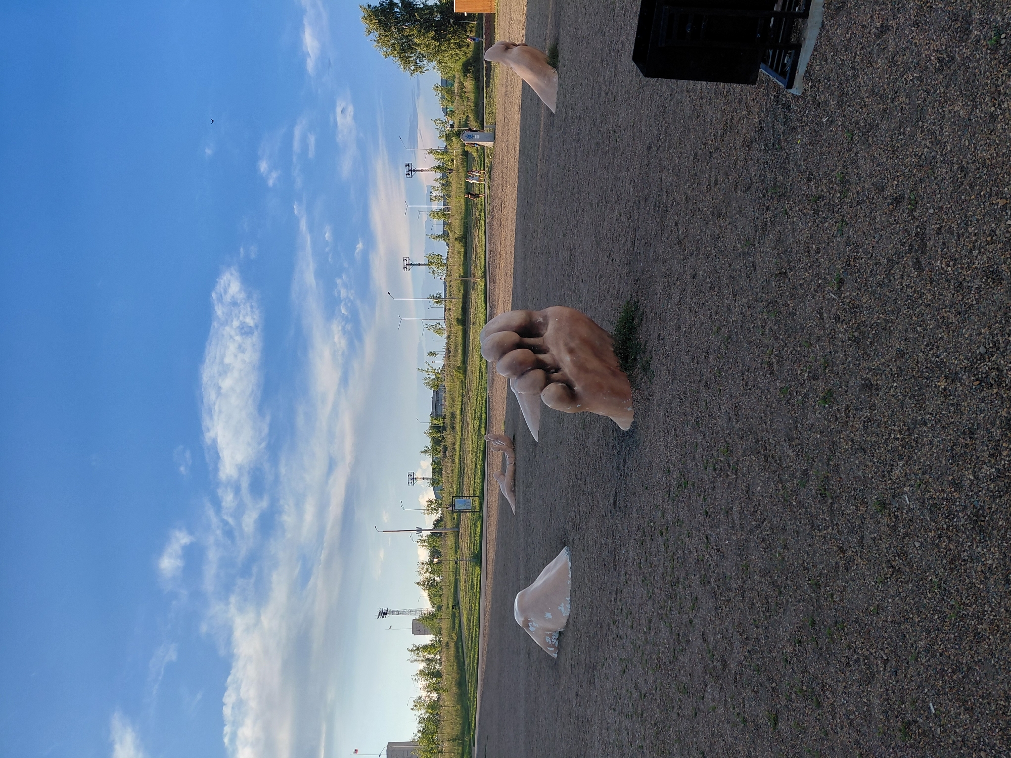 The image shows a public park in Magnitogorsk, Russia, featuring several large, stylized sculptures of human feet emerging from the ground. The sculptures are a light, flesh-like color and vary in their orientation and size. In the foreground, a large foot sculpture dominates the center of the frame, with its toes clearly defined. To the left and right, other foot or leg-like forms are partially visible, partially submerged in the gravelly ground. In the mid-ground, further sculptures are visible, as well as a group of people walking in the distance. The background reveals a grassy slope with trees, lampposts, and some industrial structures, including tall towers with flags. The sky above is blue with scattered white clouds, and a bird is visible in flight. A black waste bin is located in the lower right corner of the image. The overall environment is that of an outdoor recreational area.