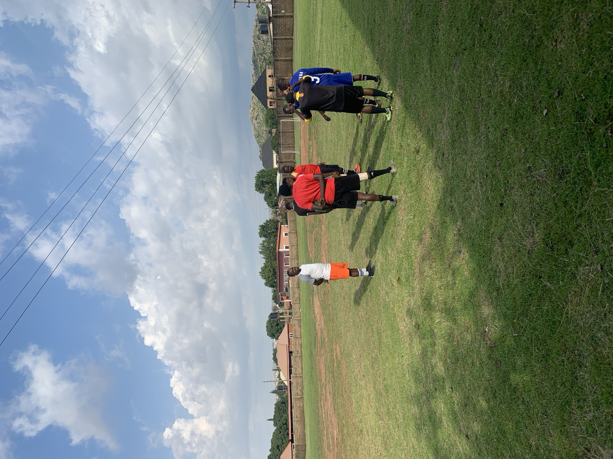 A group of young men, likely a soccer team, are gathered on a grassy field under a partly cloudy sky. They are dressed in athletic wear, including jerseys and shorts. Some appear to be engaged in a warm-up or training exercise, while others are standing and observing. The scene is set outdoors, with a fence and buildings visible in the background, suggesting an urban or semi-urban environment. The presence of power lines overhead also indicates a populated area. The overall mood seems to be one of casual activity and training. The time of day appears to be daytime, given the brightness of the sky and the shadows cast on the grass. The weather seems to be pleasant, with a mix of sunshine and clouds.