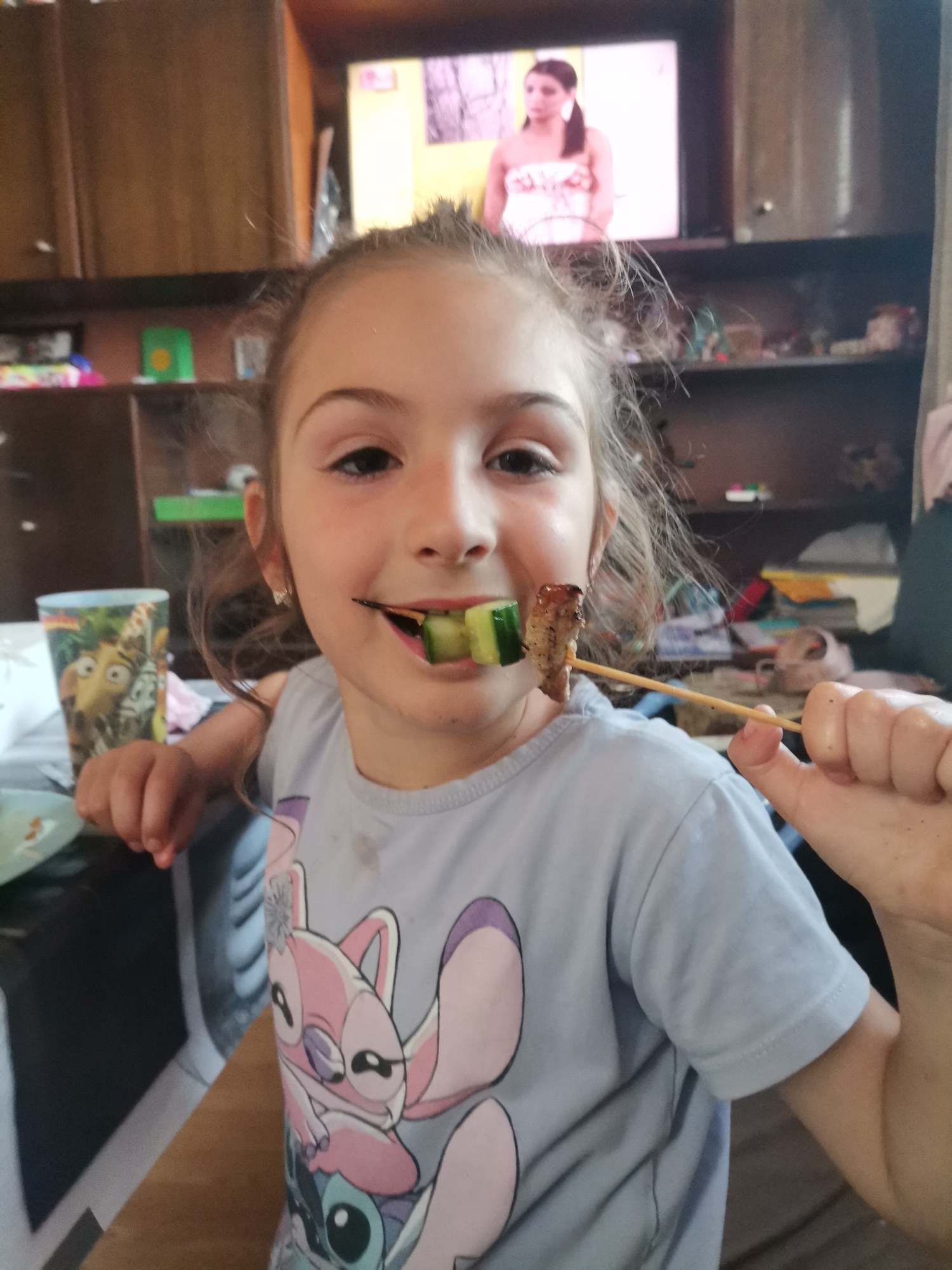 An eye-level indoor portrait captures a young girl happily eating from a skewer. The girl, with light brown hair pulled back, smiles directly at the camera. She is wearing a light blue t-shirt adorned with cartoon characters, specifically the pink alien Angel and the blue alien Stitch from Disney's Lilo & Stitch. In her right hand, she holds a wooden skewer, which contains a piece of grilled meat and two slices of green cucumber. One cucumber slice is partially in her mouth.
To her left, on a table, a clear glass cup is visible, decorated with characters from the animated movie "Madagascar," including King Julien XIII. The background shows a domestic interior, likely a home, featuring wooden cabinets and shelves containing various household items. Above these shelves, a television screen displays a program featuring a woman with dark hair, wearing a light-colored strapless top. The lighting suggests it is daytime. The girl has a small smudge on her left cheek and faint marks on her shirt, indicating a casual mealtime scene.