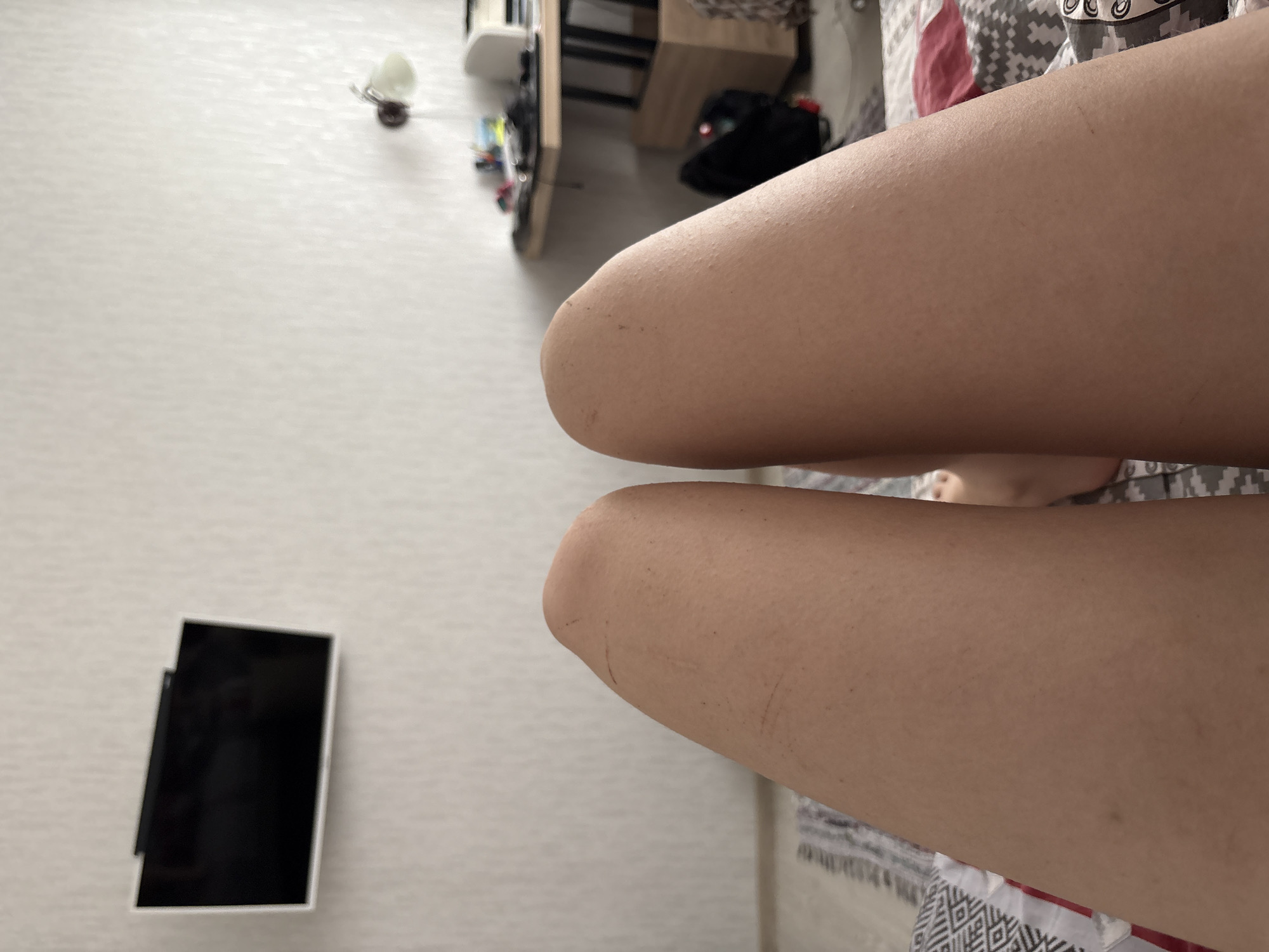 The image shows a first-person, overhead view of a person's thighs. The legs are positioned over a patterned bedsheet. A smartphone is visible on the left side of the frame, lying on a light-colored surface. In the background, there's a partially visible shelf with objects on it, and a black bag. The location is presumed to be indoors, but the city of Perm, Russia, cannot be verified from the image content alone.