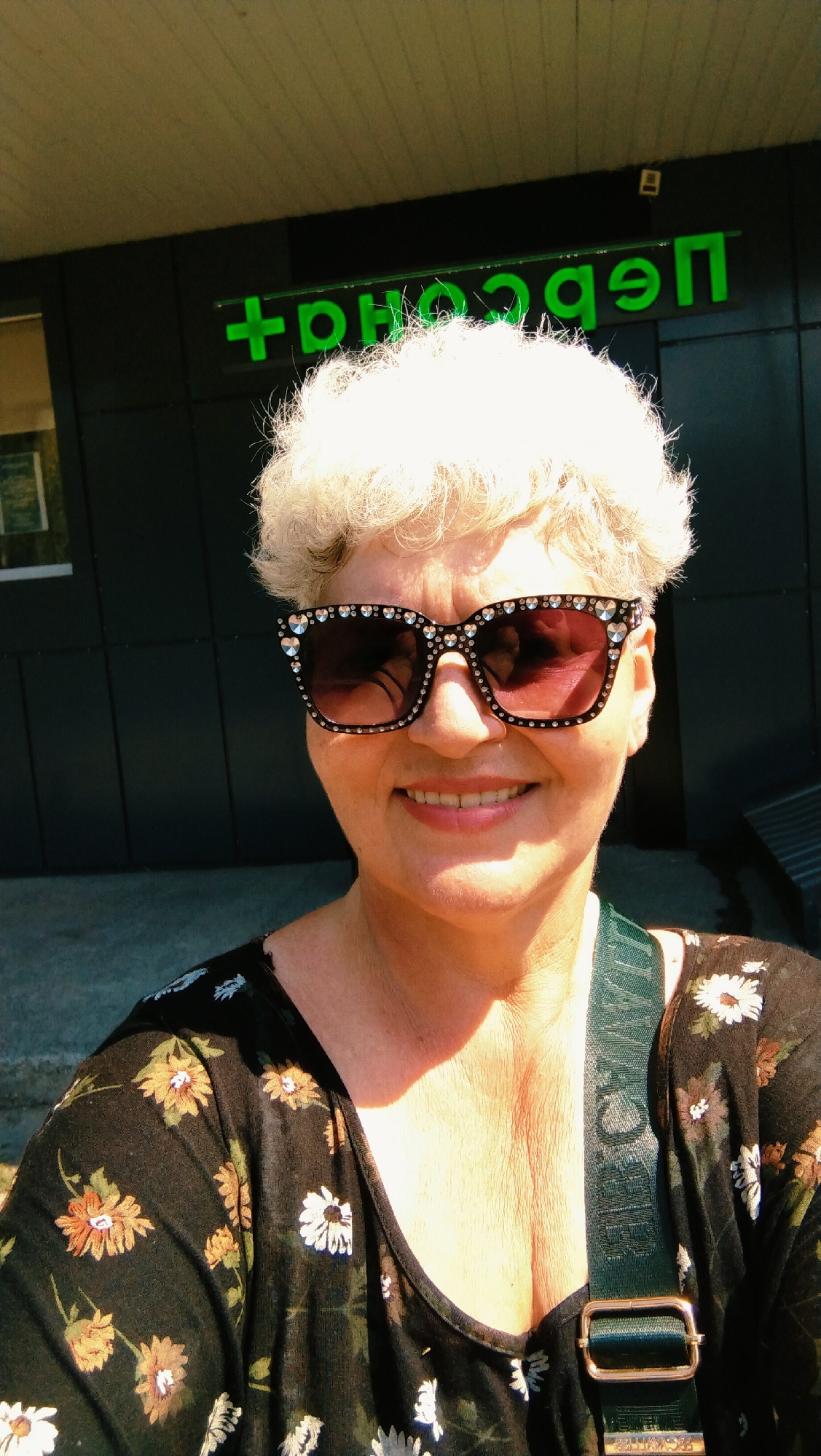 A middle-aged woman with short, white, curly hair and a floral print top smiles while wearing oversized sunglasses adorned with rhinestones. She is standing outdoors in front of a building with dark siding, where a green neon sign reads "+ Перспектива". The sun casts a shadow on her neck and chest. A dark green strap from a bag crosses her chest. The image appears to have been taken in the city of Horishni Plavni, Ukraine.