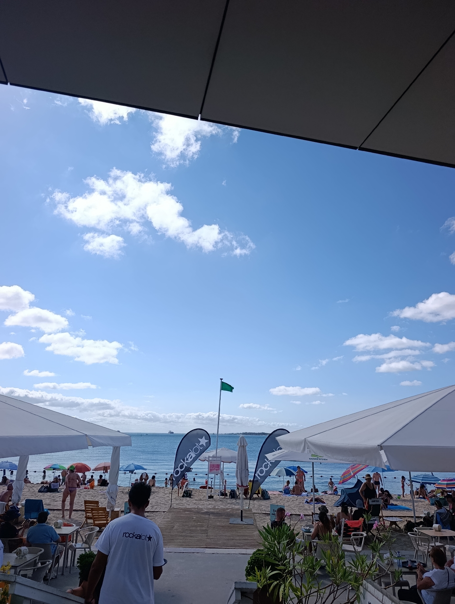 The image depicts a crowded beach scene, likely in Setúbal, Portugal, given the context. In the foreground, a man wearing a white t-shirt with a "rockalot" logo is seen from behind, facing the beach. He appears to be an employee, possibly a waiter or staff member, standing on a wooden deck. Several tables and chairs are set up on this deck, some occupied by people.
The beach itself is populated with numerous individuals sunbathing, relaxing under umbrellas, and wading in the water. Various colorful umbrellas dot the sandy shore, providing shade. The sea is visible in the background, with a few boats in the distance. The sky is blue with scattered white clouds. Two large black flags with the "rockalot" logo are prominently displayed on the beach. A green flag is also visible, possibly indicating swimming conditions. The overall atmosphere is one of a busy summer day at a popular seaside location.