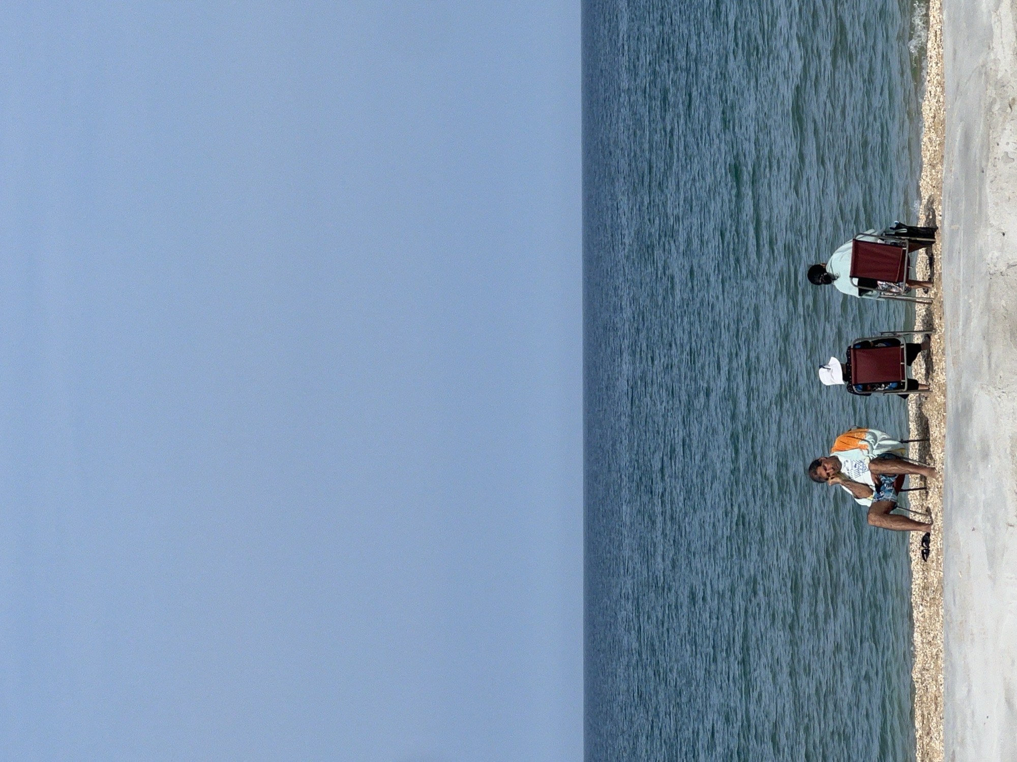 The image displays a clear, sunny daytime scene along the coast of M'Diq, Morocco. Three individuals are visible by the water's edge, engaged in what appears to be a leisurely activity.
Two people are seated on dark red foldable chairs, facing the tranquil blue sea with their backs partially or fully turned to the viewer. One person wears a light blue or green shirt, with a dark bag resting near their chair. The second seated person is partially obscured but wears a white hat. A third individual, wearing a white and orange patterned shirt and patterned shorts, is positioned closer to the viewer. They are either crouching or sitting on the edge of another red foldable chair, leaning forward with elbows on their knees, looking towards the water.
The setting consists of three distinct horizontal sections. The top portion is a vast, cloudless light blue sky. Below it is a wide expanse of deep blue sea with noticeable ripples and small waves. A narrow strip of light-colored pebbles and sand forms the shoreline, separating the water from a solid, light grey concrete or paved surface on the right side of the frame. The lighting suggests it is mid-day, with bright, direct sunlight. There is no visible text in the image.