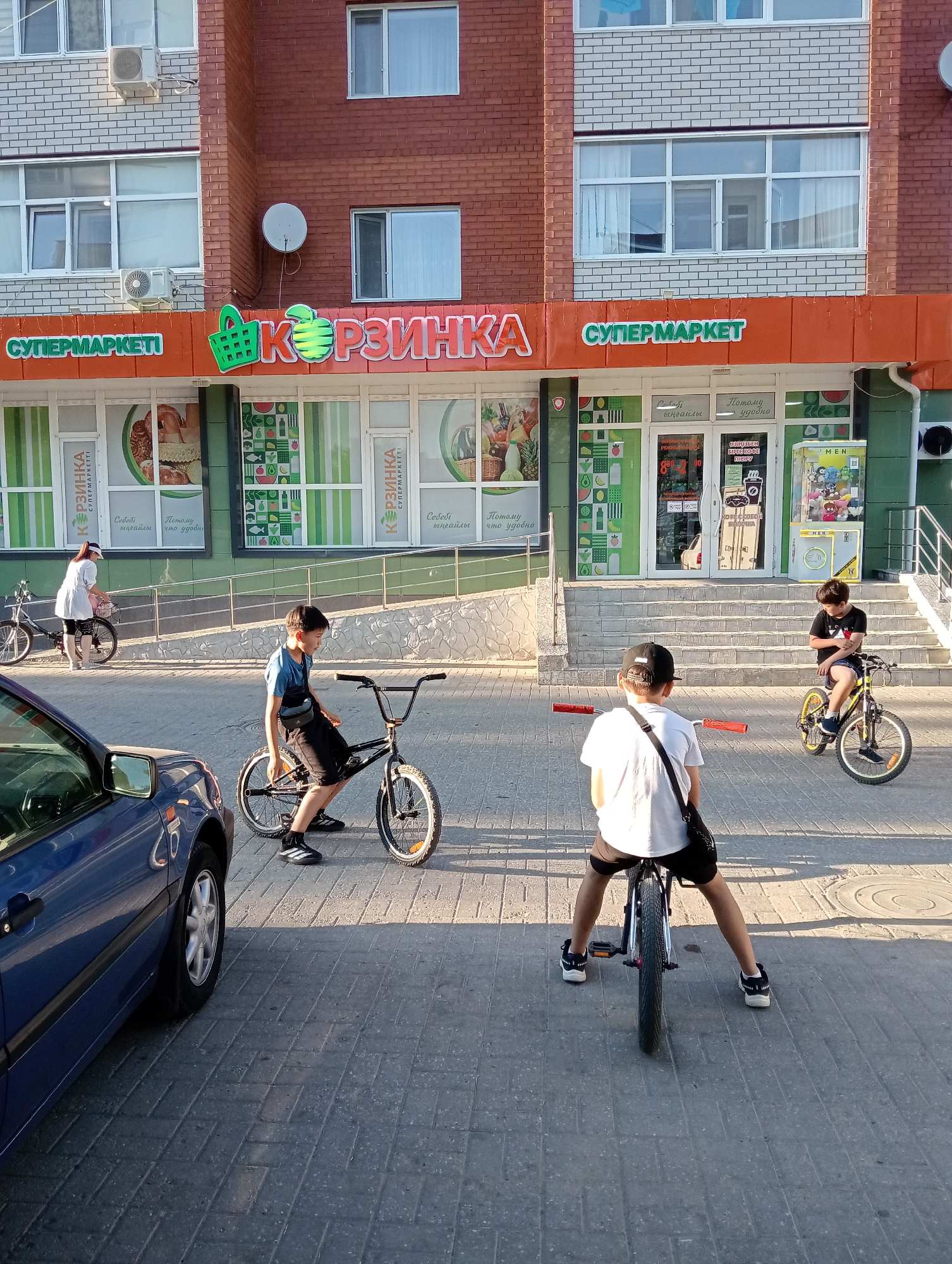 A group of four boys are seen on bicycles in front of a supermarket. The supermarket has a red and orange facade with a large sign that reads "Корзинка" (Korzinka) in large green and pink lettering. The word "СУПЕРМАРКЕТ" (Supermarket) is also displayed in green above the entrance. The building has multiple windows and air conditioning units.
In the foreground, a blue car is partially visible on the left side of the image. The ground is paved with grey interlocking bricks.
The four boys are positioned on their bicycles, appearing to be either waiting or playing. The boy on the far left is wearing a blue t-shirt and black shorts, and is on a BMX-style bicycle. The boy in the center is wearing a white t-shirt, black shorts, and a black baseball cap, and is also on a BMX-style bicycle. The boy on the right is wearing a black t-shirt and blue shorts, and is on a yellow-wheeled bicycle. The fourth boy, also wearing a black t-shirt and shorts, is on a bicycle further in the background on the right.
In the middle ground, a woman wearing a white t-shirt and dark shorts is standing next to a bicycle. The environment suggests a sunny day with dappled shadows cast by the building and surrounding objects.