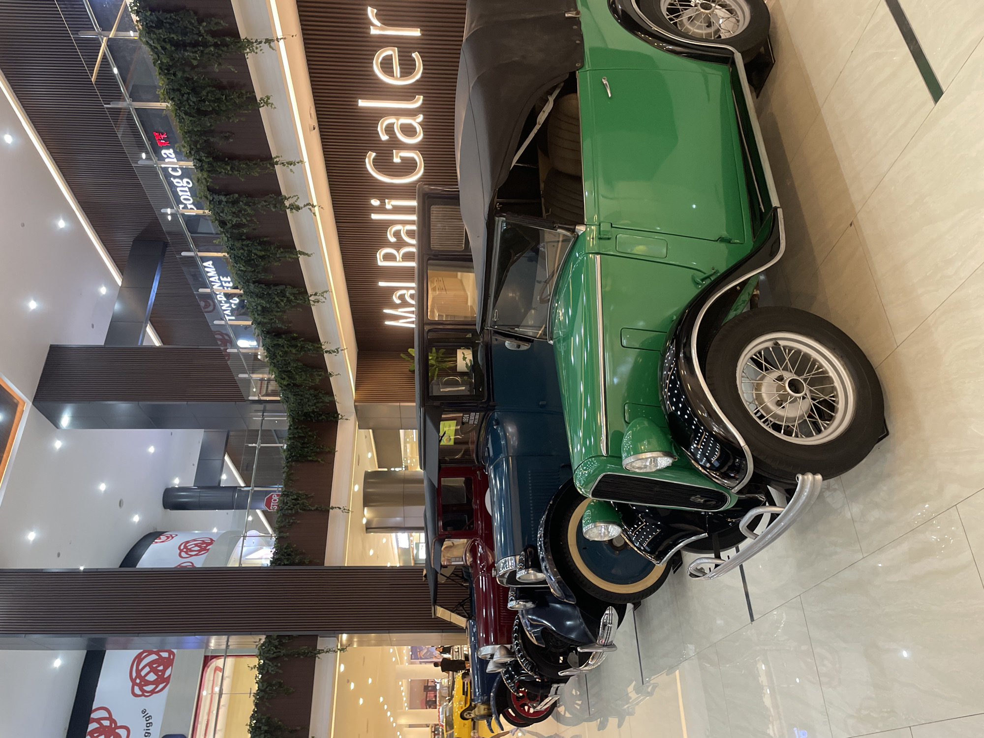 The image showcases a row of vintage cars displayed in a modern, multi-story shopping mall. A sign reading "Mal Bali Galeria" is prominent above the vehicles. The cars are in varying colors: green, blue, red, and yellow, among others. Greenery cascades from the upper floors of the mall. Shoppers are visible in the background. The mall is well-lit and has a clean, tiled floor. The photo was taken in the city of Kuta, Indonesia, at the "Mal Bali Galeria".