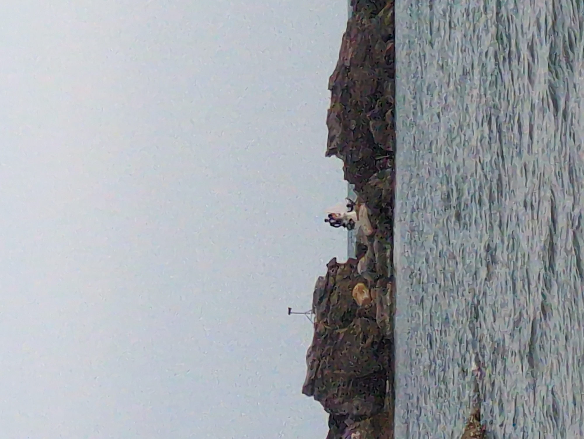 A low-resolution, rotated, and out-of-focus image depicts a coastal scene. A rocky cliff face is situated on the right side of the frame, adjacent to a body of water with a textured surface. A small structure that resembles a radio tower or antenna is visible on the cliff face. Further up the cliff, a person or people are present. The background, which takes up the majority of the frame, is a hazy grey sky. The location is likely Phú Quốc, Vietnam, based on the prompt.