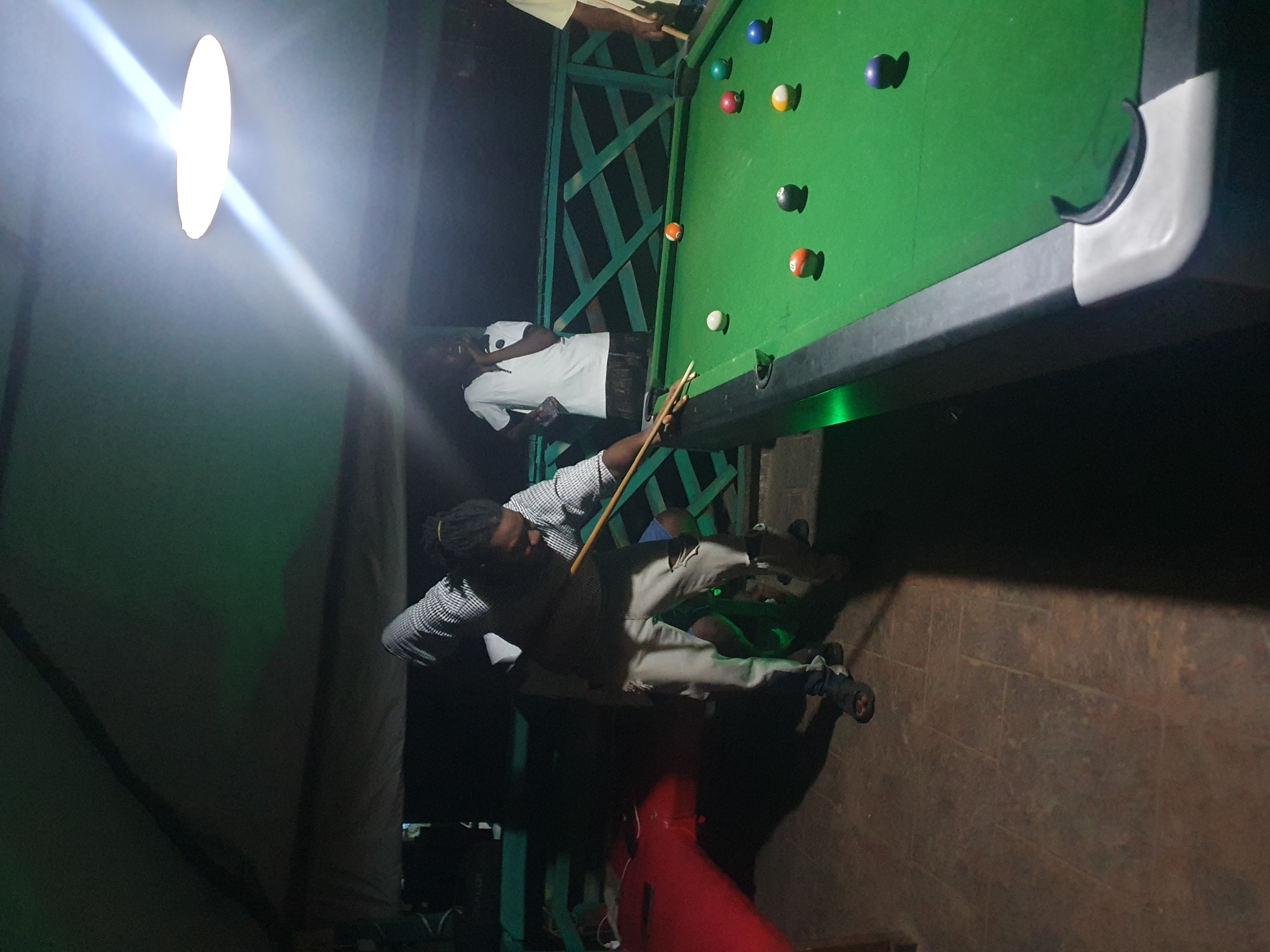 The image depicts an outdoor billiard game taking place in Lokoja, Nigeria, likely at night. A man in a checkered shirt and light-colored pants is actively playing pool, with a cue stick in his hands, aiming for a shot. He is wearing sandals. To his right, another person in a white t-shirt is observing the game, holding a phone. Further to the right, partially visible, is a person in a yellow t-shirt holding a cue stick. The billiard table is green, with several colored balls scattered across it, including orange, white, red, green, blue, black, and yellow/white striped balls. A bright overhead light fixture casts a strong illumination, creating a light beam across the scene. The surroundings suggest a casual outdoor setting, possibly a patio or veranda, with green metal railings visible in the background and a tiled floor.