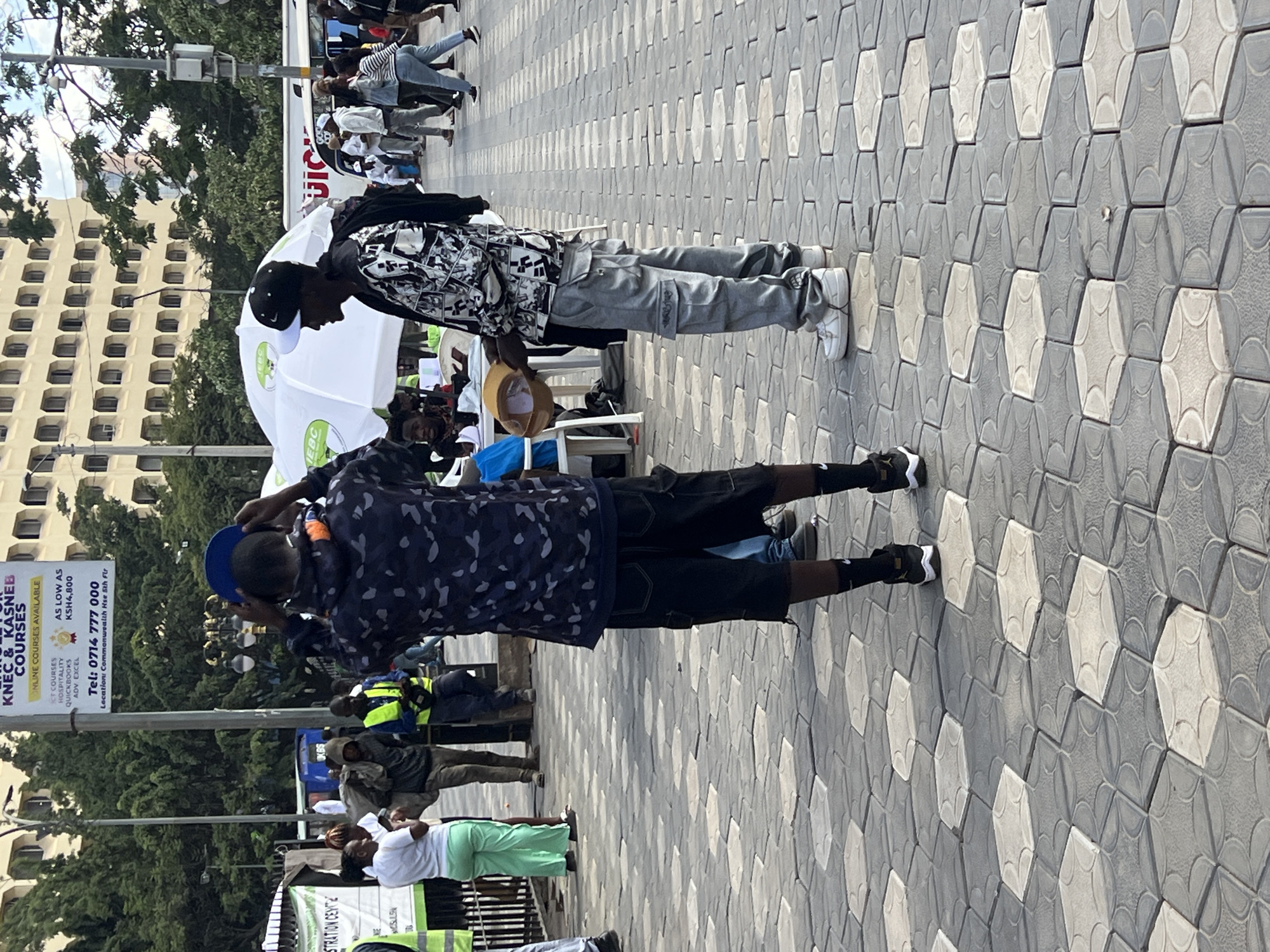 The image shows two young men standing on a sidewalk paved with a distinctive geometric pattern of gray and white hexagonal tiles. The man closest to the camera is wearing a blue baseball cap, a dark blue shirt with a light blue and white pattern, black shorts, black socks, and black and white sneakers. He appears to be adjusting his cap. The other man is wearing a black and white patterned shirt, light gray cargo pants, a black baseball cap and white sneakers, and is holding a tan hat in his hand.
In the background, there's a crowd of people, a white tent with IEBC (Independent Electoral and Boundaries Commission) logos, trees, and a tall building with a grid-like pattern of windows. A sign for KNEC & KASNEB Courses is visible, advertising online courses and giving a phone number. The location is identified as Commonwealth House, South 5th Floor. This places the scene in the city of Nairobi, Kenya.