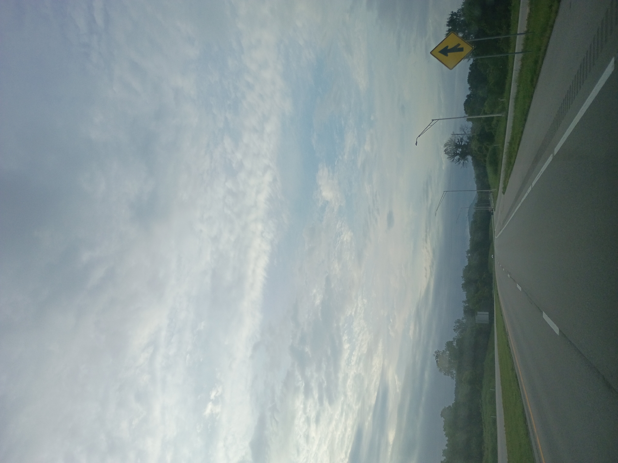 The image shows a highway scene with a cloudy sky and trees in the background. A yellow diamond-shaped sign with a black arrow pointing upwards and a line branching off to the left is visible on the right side of the road. There are streetlights along the highway. The road has clear lane markings, indicating it is a multi-lane highway. The general appearance suggests a rural or semi-rural setting outside of a major city. The location is identified as Winchester, United States.