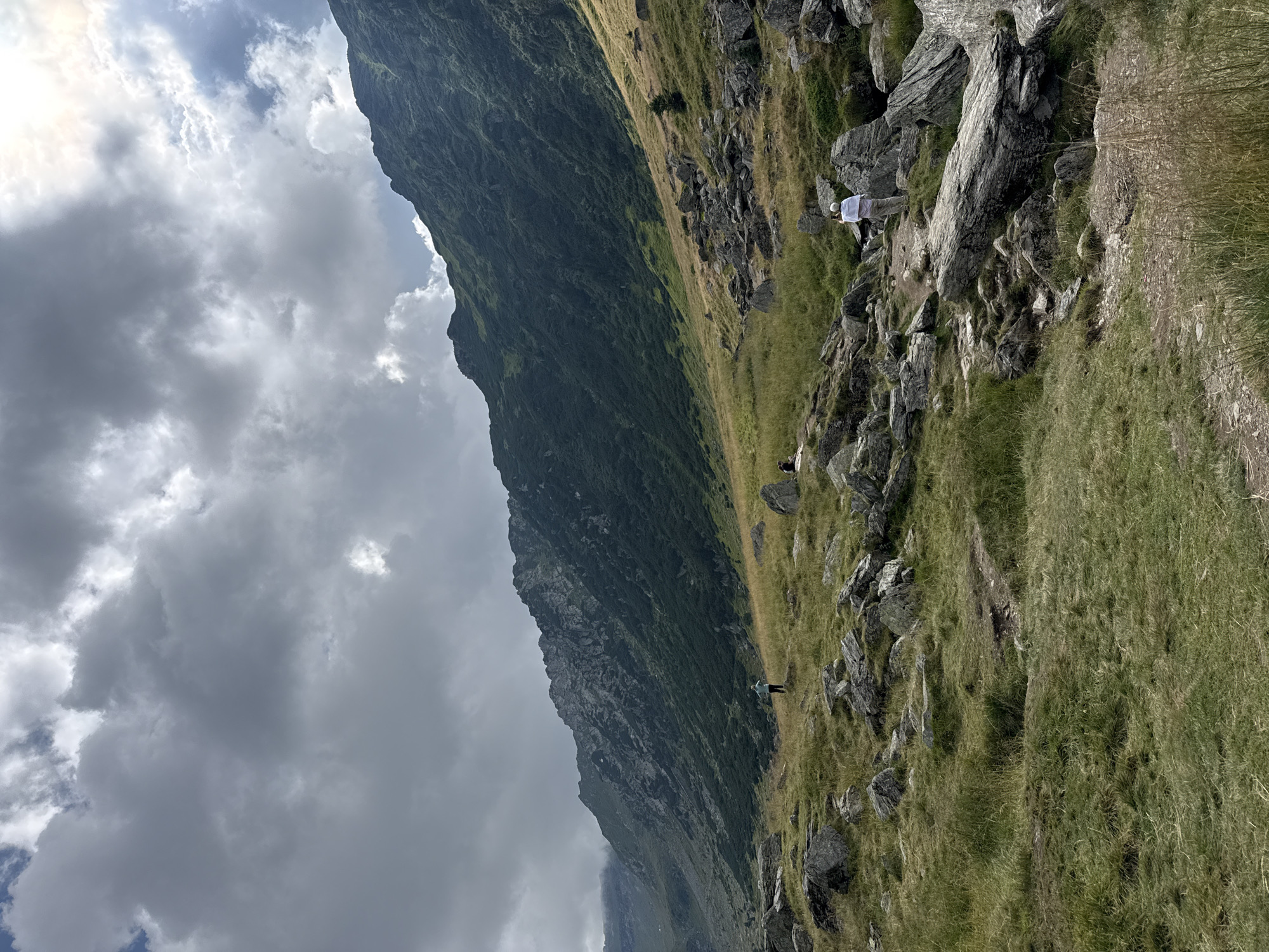 The image depicts a mountainous landscape with a cloudy sky in Cârțișoara, Romania. In the foreground, a rocky, grassy terrain with large boulders is visible. A person in a white top and light-colored pants stands on a path, facing away from the camera, appearing to observe the surroundings. Further in the mid-ground, another person in a teal top stands on a grassy slope, also facing away. In the background, a steep, verdant mountain slope rises, covered in dense vegetation. The sky is filled with dramatic, dark clouds, with patches of sunlight breaking through.