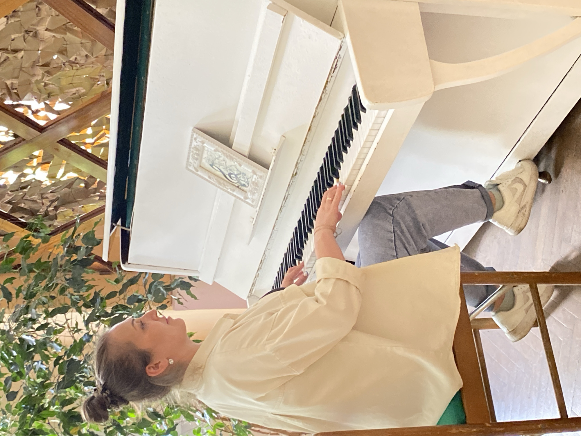 An indoor scene captured in Марковский, Russia, shows a woman playing a white upright piano. The image is oriented vertically but depicts the woman seated at the piano, with her profile visible on the left side of the frame. She has light hair pulled back into a bun and wears a pearl stud earring. Her attire consists of a light-colored, possibly cream or off-white, long-sleeved button-up shirt and light blue or grey trousers. Her right hand is positioned on the piano keys, indicating she is actively playing. Underneath the piano, her feet are visible, clad in white sneakers.
The white piano is a prominent feature, with its black and white keys clearly seen. A small, ornate framed object, possibly a picture or decorative plaque, rests on the piano's top surface. The woman is seated on a wooden chair with a light-colored cushion.
The background suggests a vibrant and somewhat eclectic interior. To the left of the woman and behind the piano, lush green leafy plants are visible, adding a touch of nature to the setting. Further in the background, there are architectural elements featuring wooden beams and a textured, reflective ceiling or wall section, which appears to be made of a crinkled, metallic material, possibly foil, reflecting light and creating bright spots. The lighting suggests it is daytime, with natural light illuminating the space. The woman's expression appears calm and focused, as she looks slightly upwards, likely concentrating on her music. No discernible text is visible in the image.