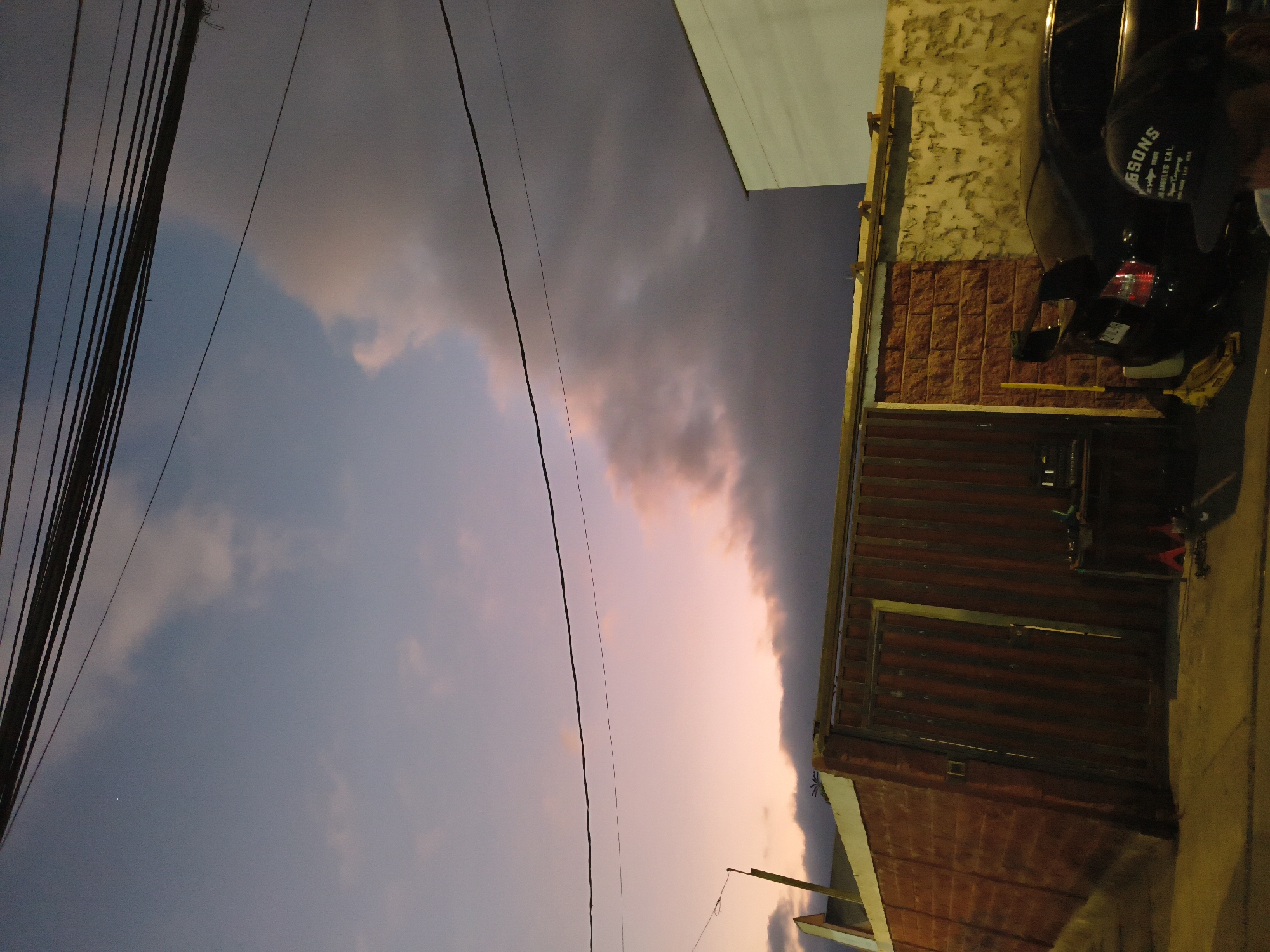 The image, rotated 90 degrees, depicts a street scene in Arica, Chile at dusk. The sky transitions from blue to pink and grey, with electrical wires prominent in the upper left. A building is visible on the right side, showing a brick facade at the top and a corrugated metal section below. A black motorcycle is parked in front of the building, partially obscuring a sign on the side that says "GSONS". A sidewalk is visible along the bottom edge of the image, illuminated by street lighting.