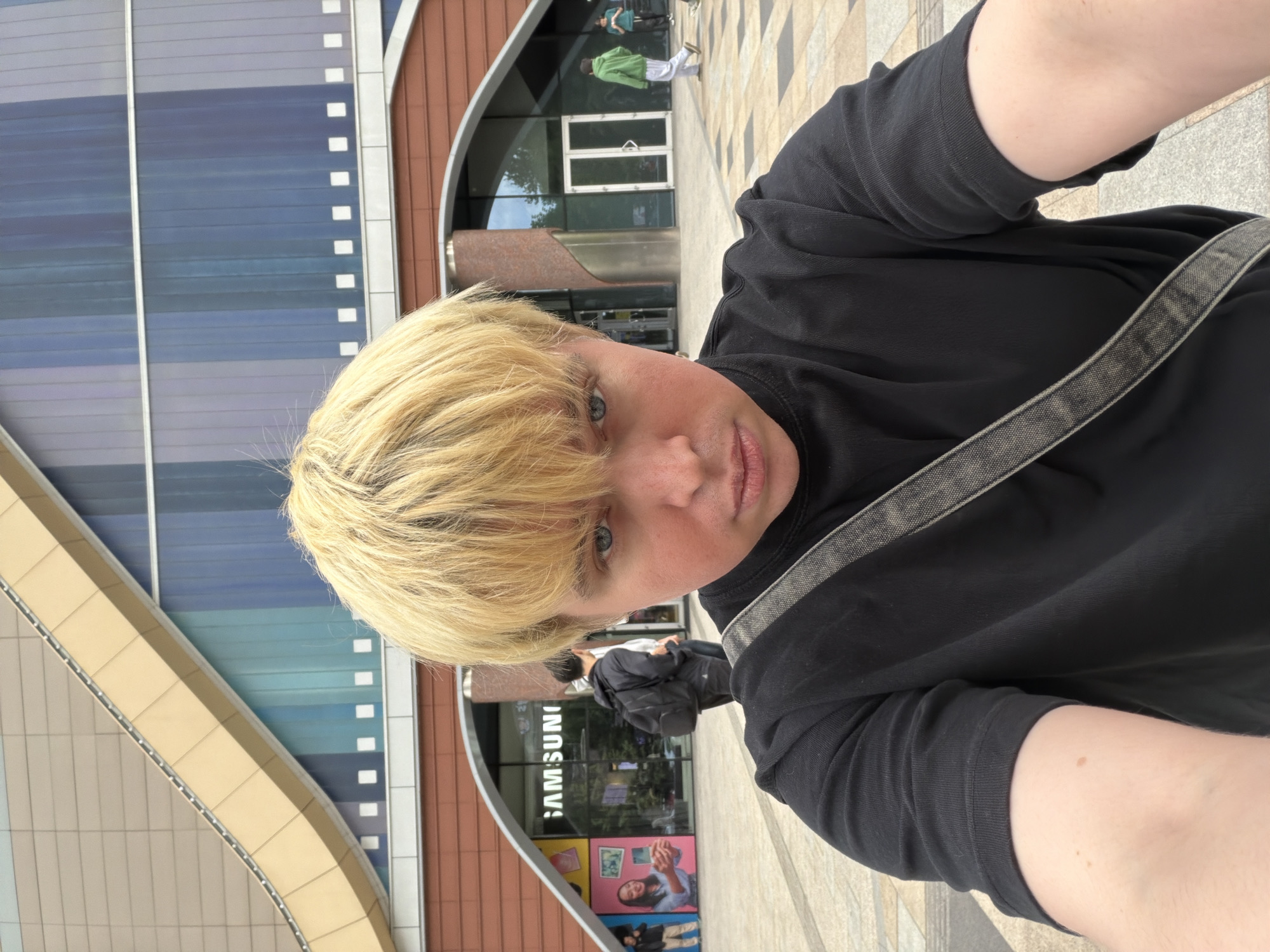 The image depicts a selfie taken outdoors. The subject, positioned in the foreground, has fair skin, blonde hair, and is wearing a black top with a grey strap. Behind the subject, a building with a curved facade dominates the background. The building's exterior features blue and brown panels, interspersed with white accents. The reflection on the glass shows the word "SAMSUNG". Below that are posters of photos. The environment appears to be a modern urban area with a tiled sidewalk. Other people are barely visible in the background.