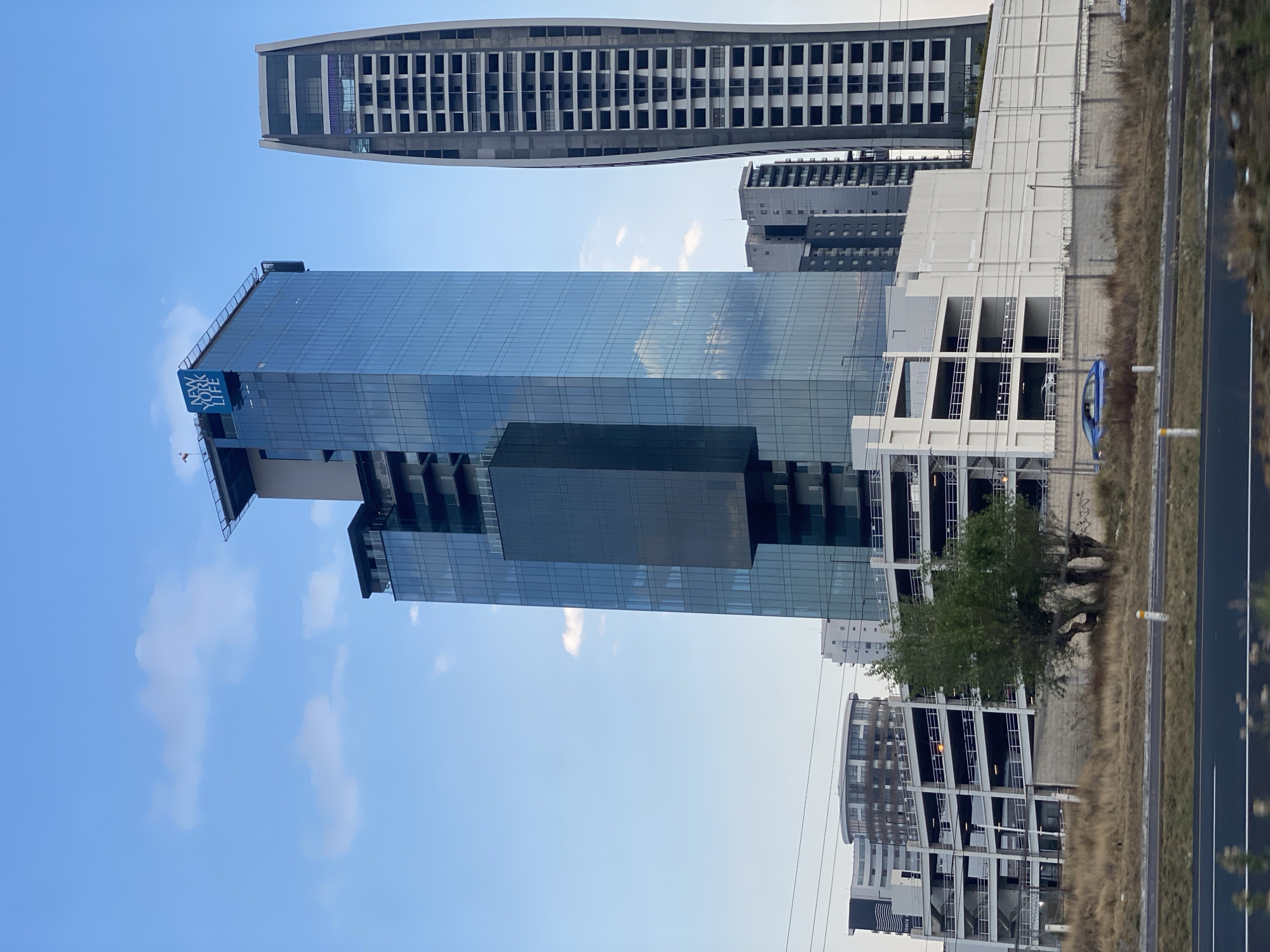 The image shows a view in San Bernardino Tlaxcalancingo, Mexico, featuring several modern buildings under a blue sky with scattered clouds. Prominently displayed is a tall, reflective glass building with the 'New York Life' logo visible on its upper section. A taller, unfinished-looking building with repetitive window patterns stands slightly behind and to the left of the glass structure. To the right of the main glass building, there's a multi-level parking garage structure and other buildings that suggest a commercial or residential zone. A tree is positioned near the parking structure, and a road with a median or curb is visible in the foreground, with dry vegetation along its edge.