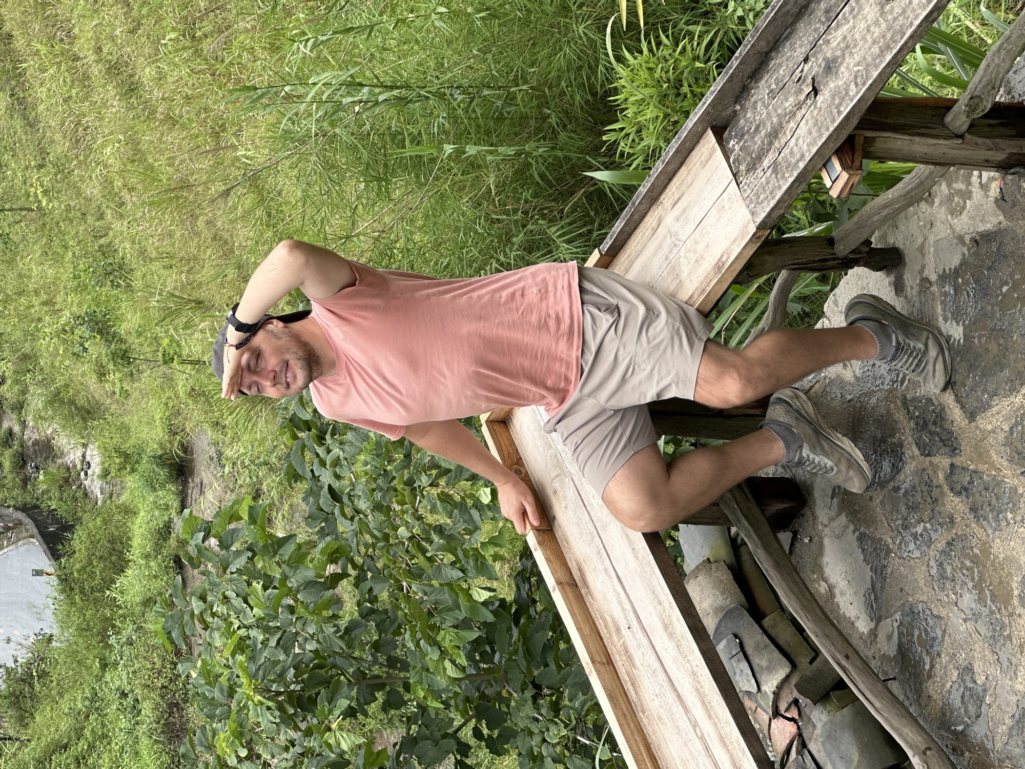 A person is standing on a weathered wooden structure in Yên Minh Commune, Vietnam, likely a viewing platform or elevated walkway. The person has a beard and dark hair, wearing a pink short-sleeved shirt, tan shorts, and gray sneakers. Their right hand is raised to their forehead, shielding their eyes. The background includes dense green vegetation, including trees and tall grasses, suggesting a natural environment. The wooden structure is built over a textured stone or tile surface.