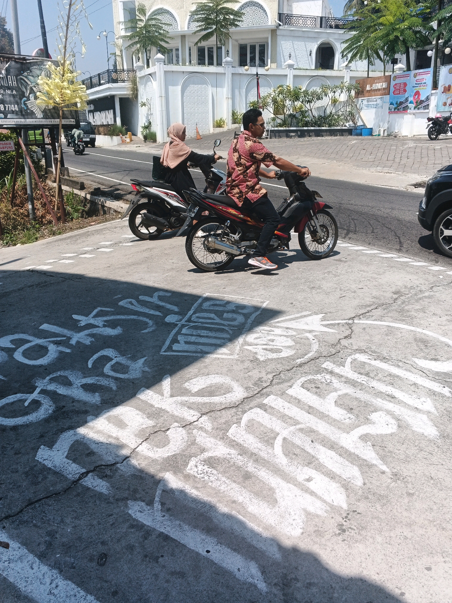 The image depicts a street scene in Pakopen, Indonesia, with a focus on a busy intersection where two individuals are riding motorcycles. In the foreground, chalk markings are visible on the asphalt, with what appears to be text and directional arrows painted in white. A light-colored shadow falls across the lower portion of the image, partially obscuring the markings.
On the right side of the frame, a man wearing a red and white patterned shirt and dark pants rides a red motorcycle. He is looking forward and appears to be in motion. On the left side, a person wearing a light brown hijab and a dark garment rides a motorcycle. Both motorcycles are positioned on the road, with lane markings visible behind them.
In the background, a white building with arched windows and ornate architectural details stands prominently. Palm trees and other greenery are present in front of the building. Several billboards are displayed on the building's facade. A black car is partially visible on the far right of the image, indicating traffic flow. The sky is clear and bright, suggesting a sunny day. The overall environment appears to be an urban or semi-urban setting.