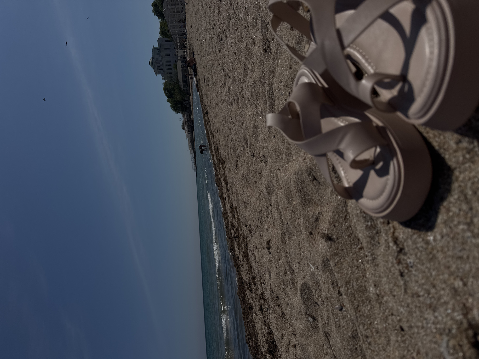 The image depicts a beach scene in Yevpatoriya, Ukraine. A pair of beige sandals sit on the sandy beach. The sea is visible in the background, along with a building along the shoreline. The sky is blue with some birds visible in the sky. There are a few people in the water.
