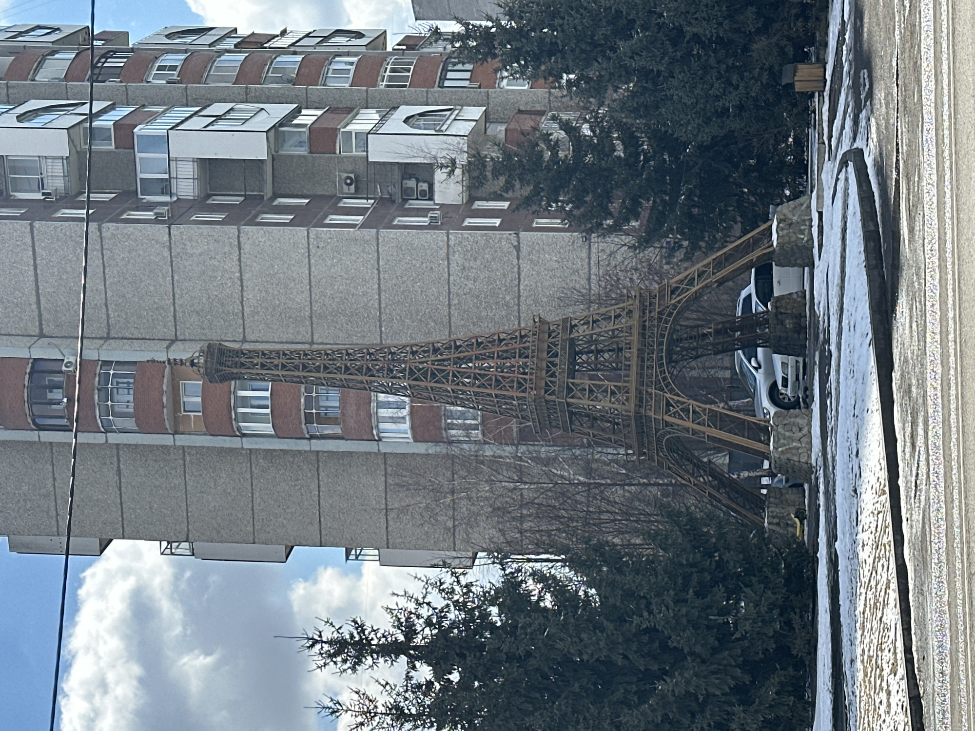 The image shows a replica of the Eiffel Tower in front of a tall building in what appears to be an urban environment. The building has a mix of concrete and brick or tile facade, with several rows of windows and balconies. The sky is visible in the background with some clouds. There are several trees and other greenery around the Eiffel Tower. A white car is parked near the Tower. The location is identified as Krasnoyarsk, Russia.
