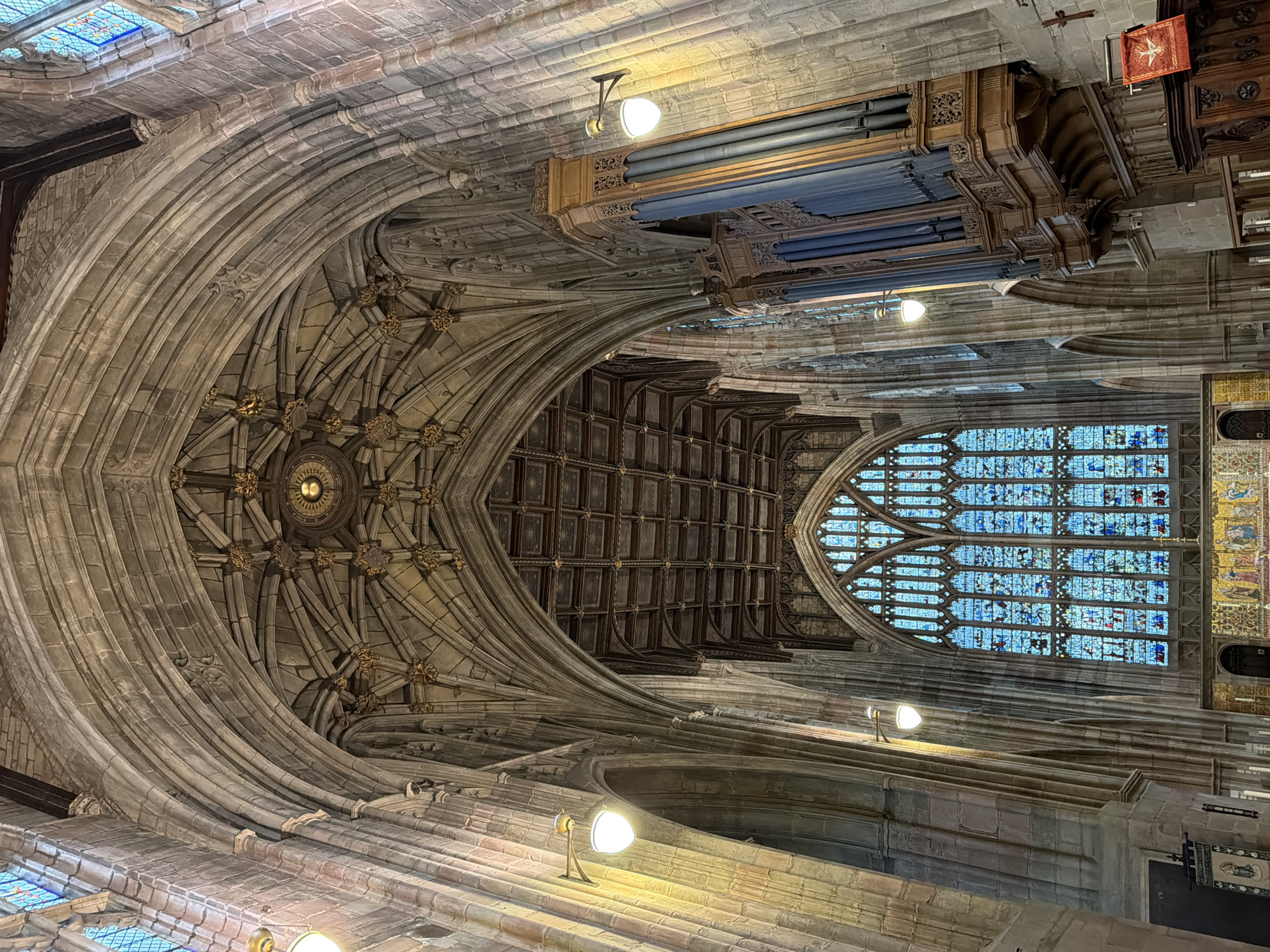 The image depicts the interior of a grand, gothic-style building, possibly a cathedral or church, in Malvern Hills, United Kingdom. The architectural style is characterized by vaulted ceilings, ribbed arches, and stained glass windows. The primary focus is the elaborate ceiling, adorned with intricate patterns and decorative elements, including a central, ornate circular design. A large pipe organ is visible near the top of the frame, adding to the setting's religious character. The stained glass windows filter light into the space, creating a colorful illumination within the structure. The scene is devoid of people and focuses on the structural and decorative aspects of the interior architecture. The image has been rotated by 90 degrees to the right.