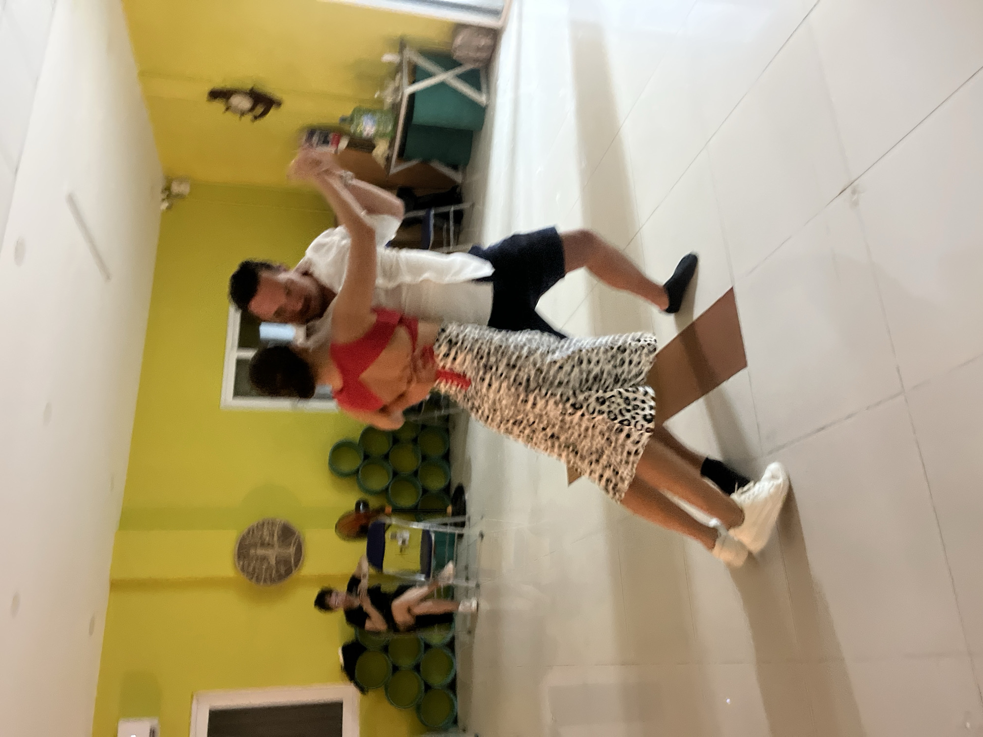 The image depicts an indoor dance studio with bright yellow walls, located in the city of Đà Nẵng, Vietnam. In the foreground, a couple is engaged in dancing, with the woman wearing a leopard print skirt and the man in a white shirt and dark shorts. The dance appears dynamic, with their bodies positioned in a pose of movement. In the background, a woman is seated on a chair, looking at her phone, with a fan and stacked circular objects behind her. The floor is tiled and reflective, suggesting a clean and well-maintained space. Several objects, including a clock and what appear to be water bottles, are visible on a shelf in the background.