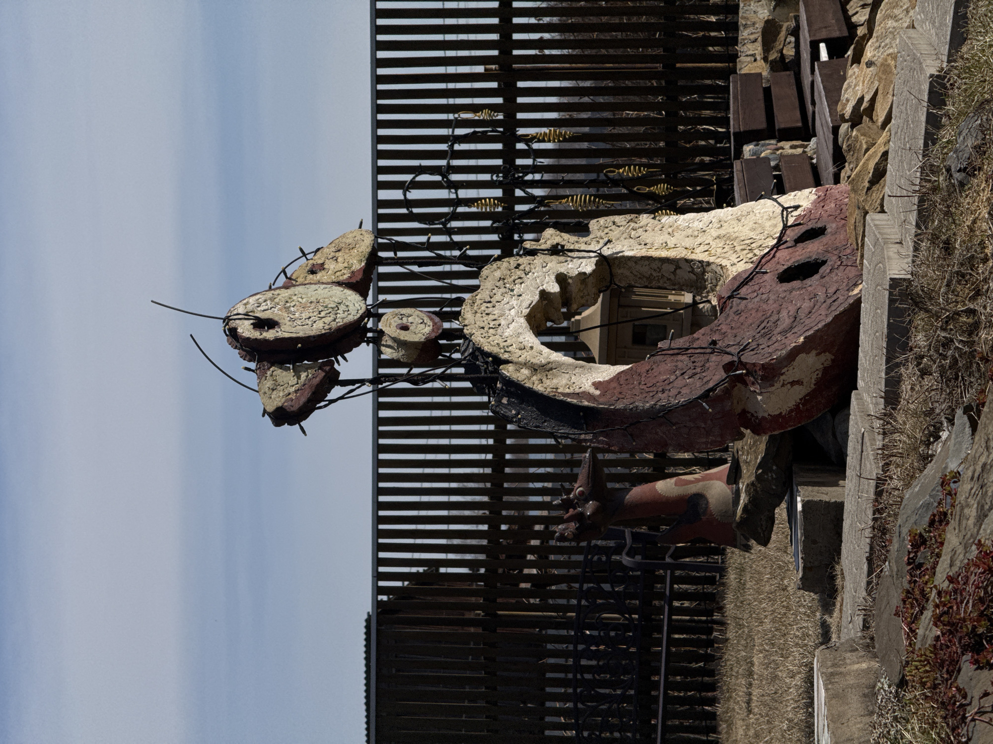The image depicts an outdoor art installation in what appears to be the city of Sukhobuzimskoye, Russia. The foreground features a sculptural arrangement comprised of painted and textured elements, including circular and ring-shaped components connected by wire. The color palette includes reddish-brown, white, and off-white hues.
A dark, slatted structure serves as a backdrop, adorned with small, metallic fish figures. Behind the sculpture, a pile of stones and wooden planks is visible, along with some vegetation growing on the edge. The sky is visible in the upper left corner.