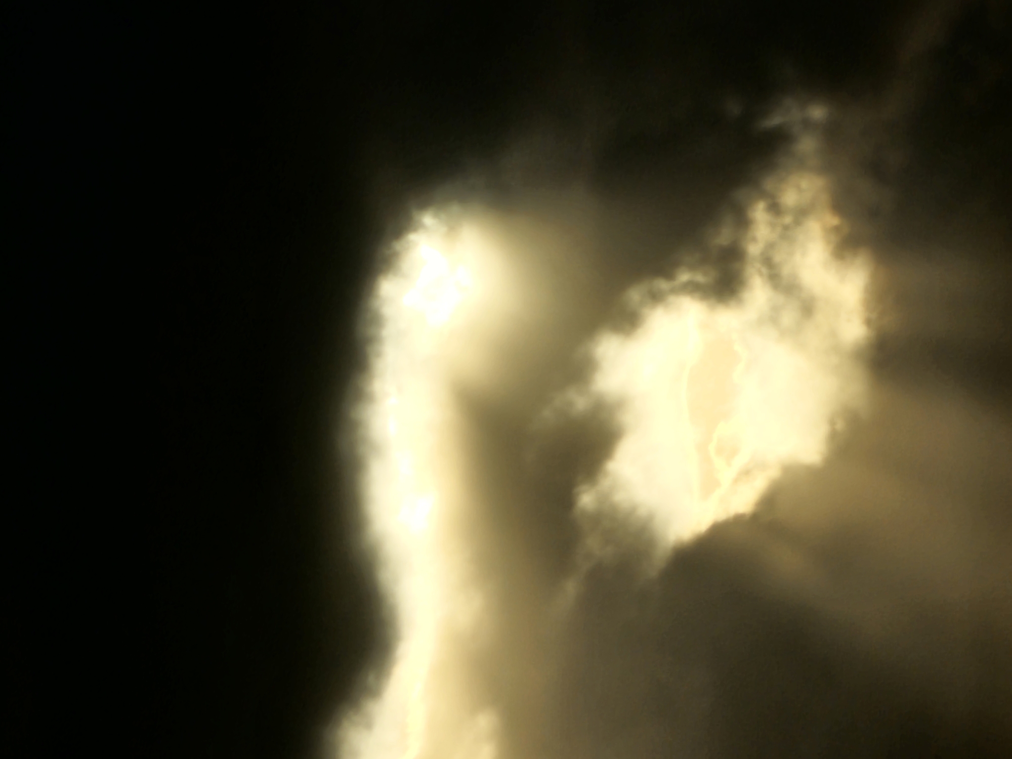 The image depicts a dramatic sky filled with dark, ominous clouds pierced by shafts of brilliant, golden sunlight. The light breaks through the clouds in irregular patterns, creating a contrast between light and shadow. The scene is abstract, focusing on the interplay of light and atmospheric elements rather than specific objects or human activity. There is no identifiable location within the city of Denpasar, Indonesia. The overall impression is one of natural, turbulent beauty.