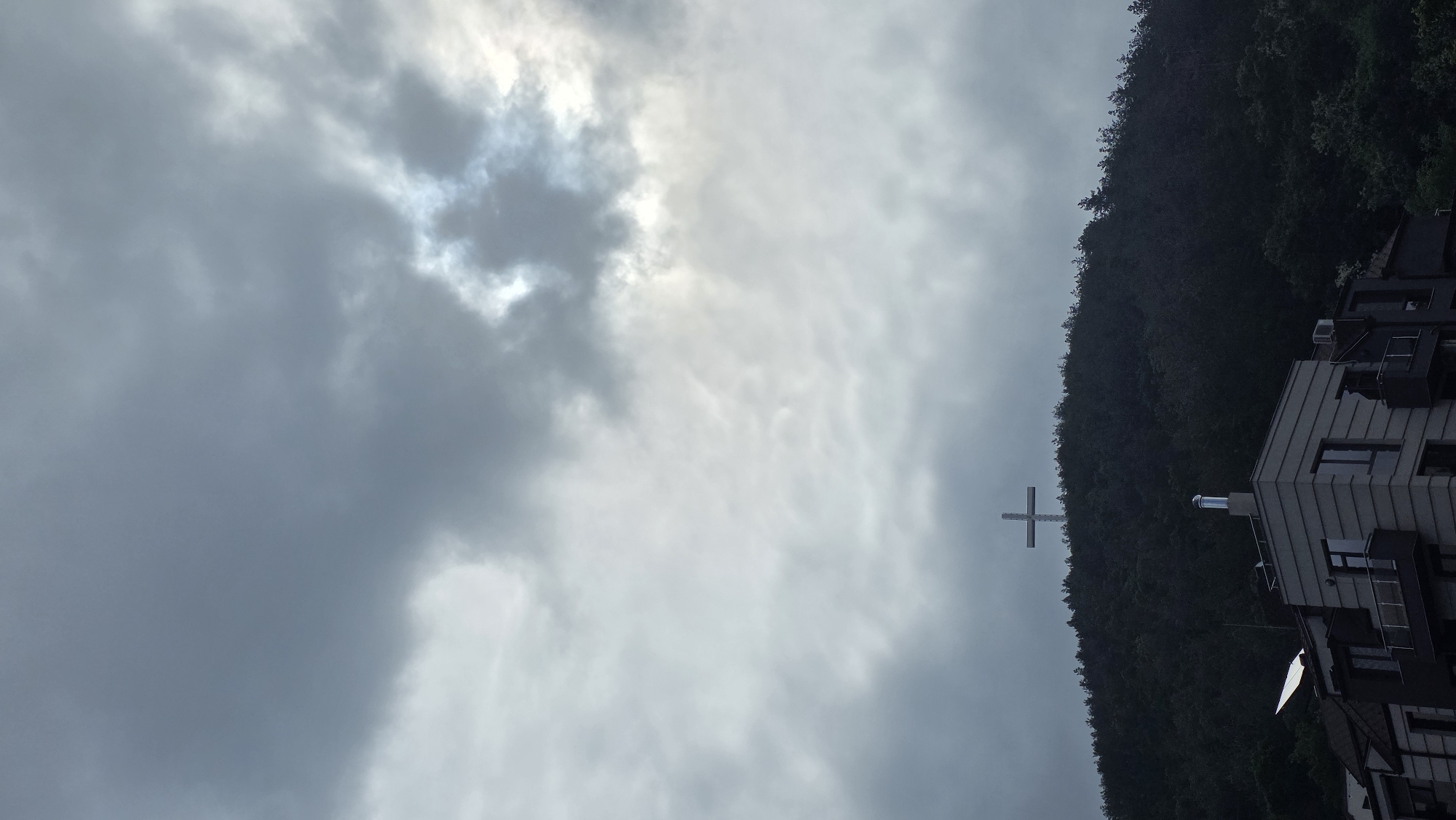 A low-angle shot shows a cloudy sky dominating the frame, with a sliver of bright light peeking through the clouds in the upper left corner. The right side of the image features a wooded hillside descending towards a building complex. A tall, metal cross stands prominently against the backdrop of the trees, near the building. The buildings appear modern and multi-storied, with dark roofing and window accents. The entire scene is enveloped in a muted, overcast light. The location appears to be Blagoevgrad, Bulgaria.