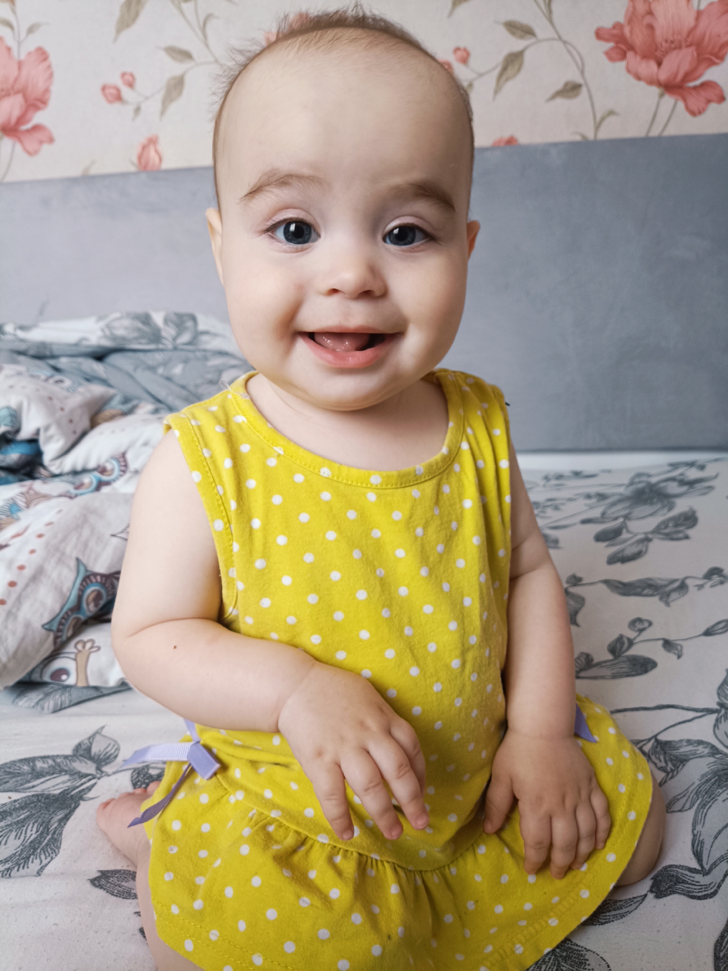 A close-up, medium shot captures a young Caucasian baby sitting on a bed. The baby, with fair skin and light brown hair, is wearing a yellow sleeveless dress with white polka dots. A light purple bow is tied on the left side of the dress. The baby's hands are resting on its lap. The background consists of a bed with patterned bedding and a wall with floral wallpaper. The baby's expression is one of joy, with eyes wide open and a broad smile revealing its teeth and tongue. This image was taken in Noviy, Russia.