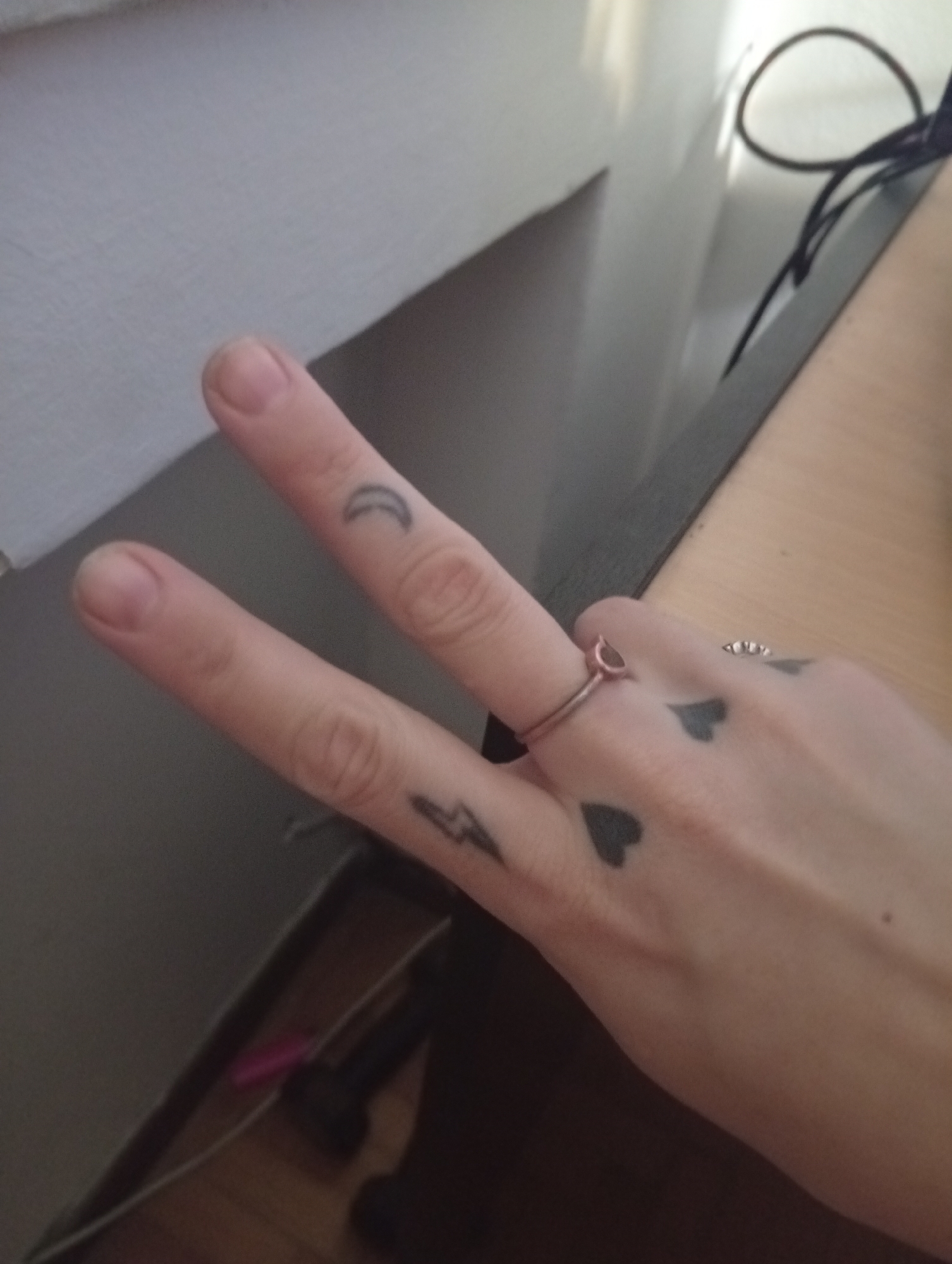 A person's hand is positioned in the foreground, making a peace sign with the index and middle fingers. The hand has multiple small tattoos, including a crescent moon on the index finger, a lightning bolt on the middle finger, and several hearts on the back of the hand. The hand is also adorned with two rings, one on the ring finger and another on the pinky. The background consists of a white wall and a desk with a dark edge. A power cord is visible on the desk surface. The location is indeterminate.