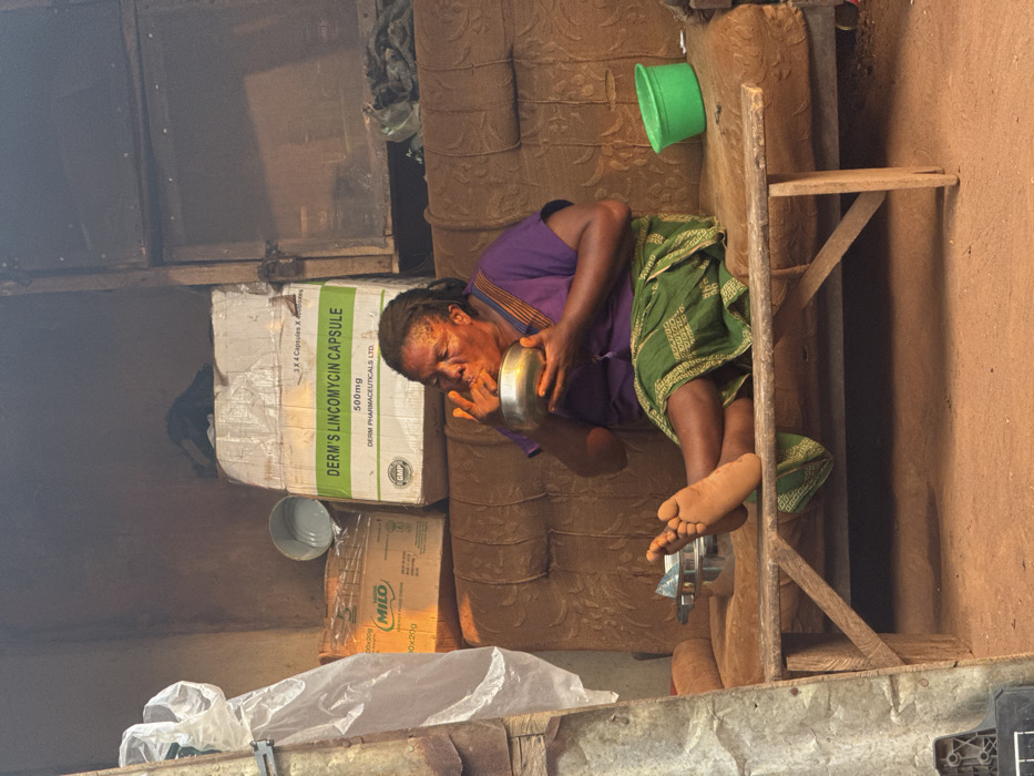 An older woman in a purple shirt and green patterned skirt sits on a wooden stool, holding a silver bowl to her lips. The scene appears to be indoors, with various boxes stacked behind her. One box is labeled "DERM'S LINCOMYCIN CAPSULE 500mg," and another is labeled "MILO." A green bucket hangs on the wall nearby. The background shows simple construction.