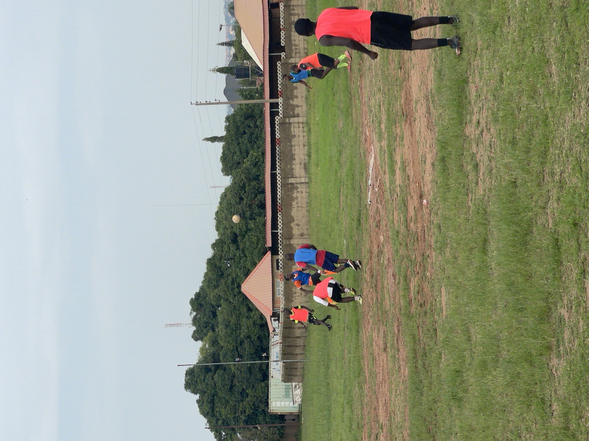In Jos, Nigeria, on an overcast day, a group of men in athletic wear are engaged in a soccer practice on a dirt and grass field. The men are wearing brightly colored training bibs over their shirts, with some in orange and others in blue and red. The action captures a moment of play, with a soccer ball in mid-air, being kicked towards the goal. The background reveals a mix of urban and natural elements, including a cluster of tall, lush green trees, a long stone wall with a decorative top, a shipping container, and distant buildings with red roofs. A radio tower is visible in the far background. The overall scene depicts an active sports training session in a typical outdoor environment.